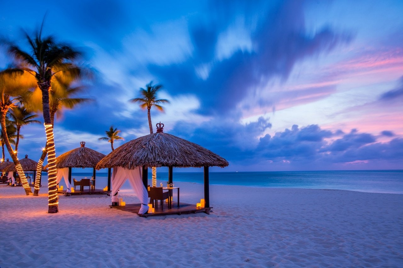 A line of grass-thatched gazebos with a table and two chairs inside face the sea during dusk. Behind are a row of palm trees intertwined with fairy lights; caribbean sea turtles