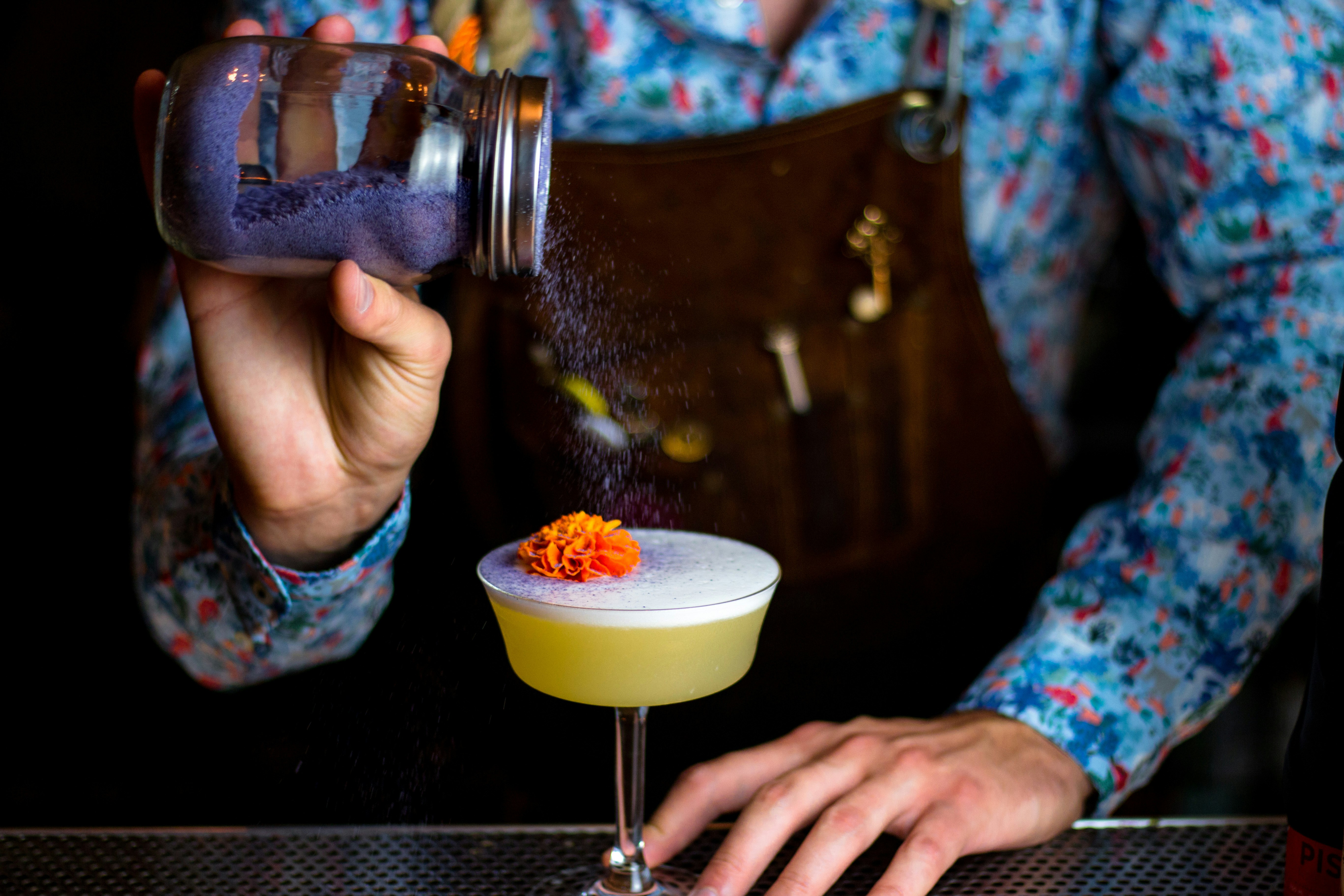 A bartender sprinkles purple garnish powder on the top of a frothy cocktail with an orange flower at the top
