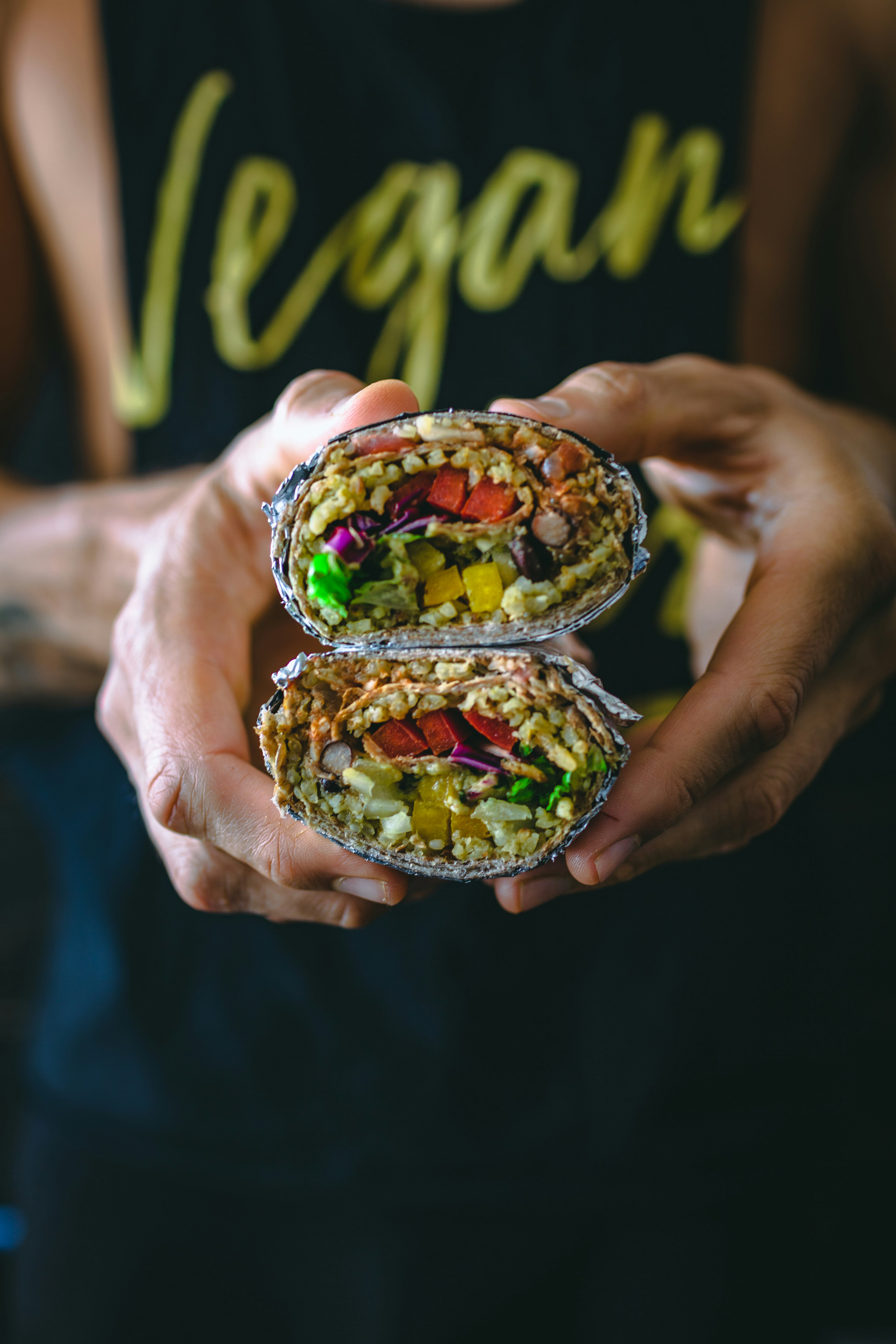 A man wearing a t-shirt that says 'vegan' holds a burrito filled with vegetables towards the camera 