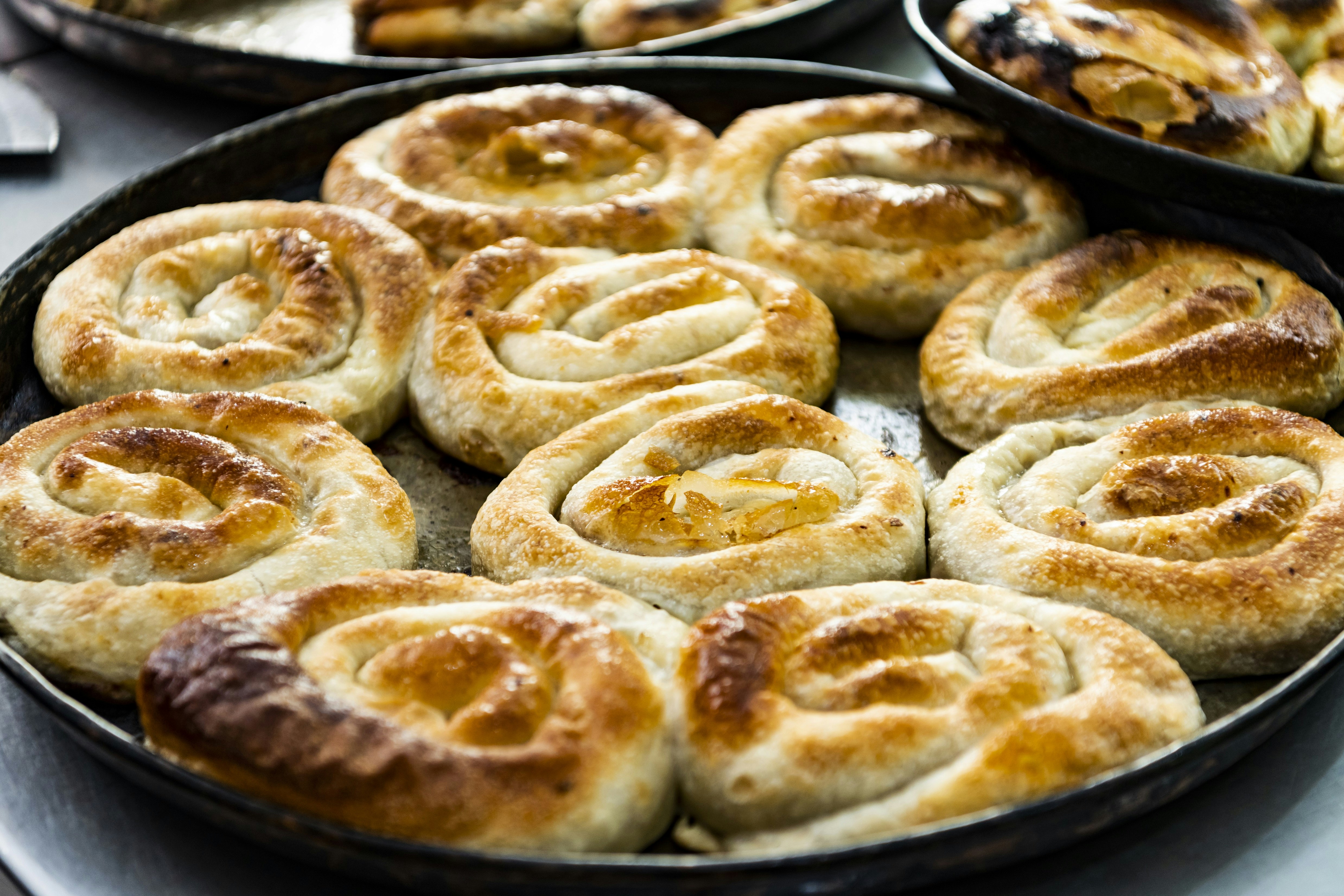 A dozen burek pies cooling on a large black plate. The pies are spirals resembling a snail shell, made from pastry and containing various ingredients like meat and vegetables.
