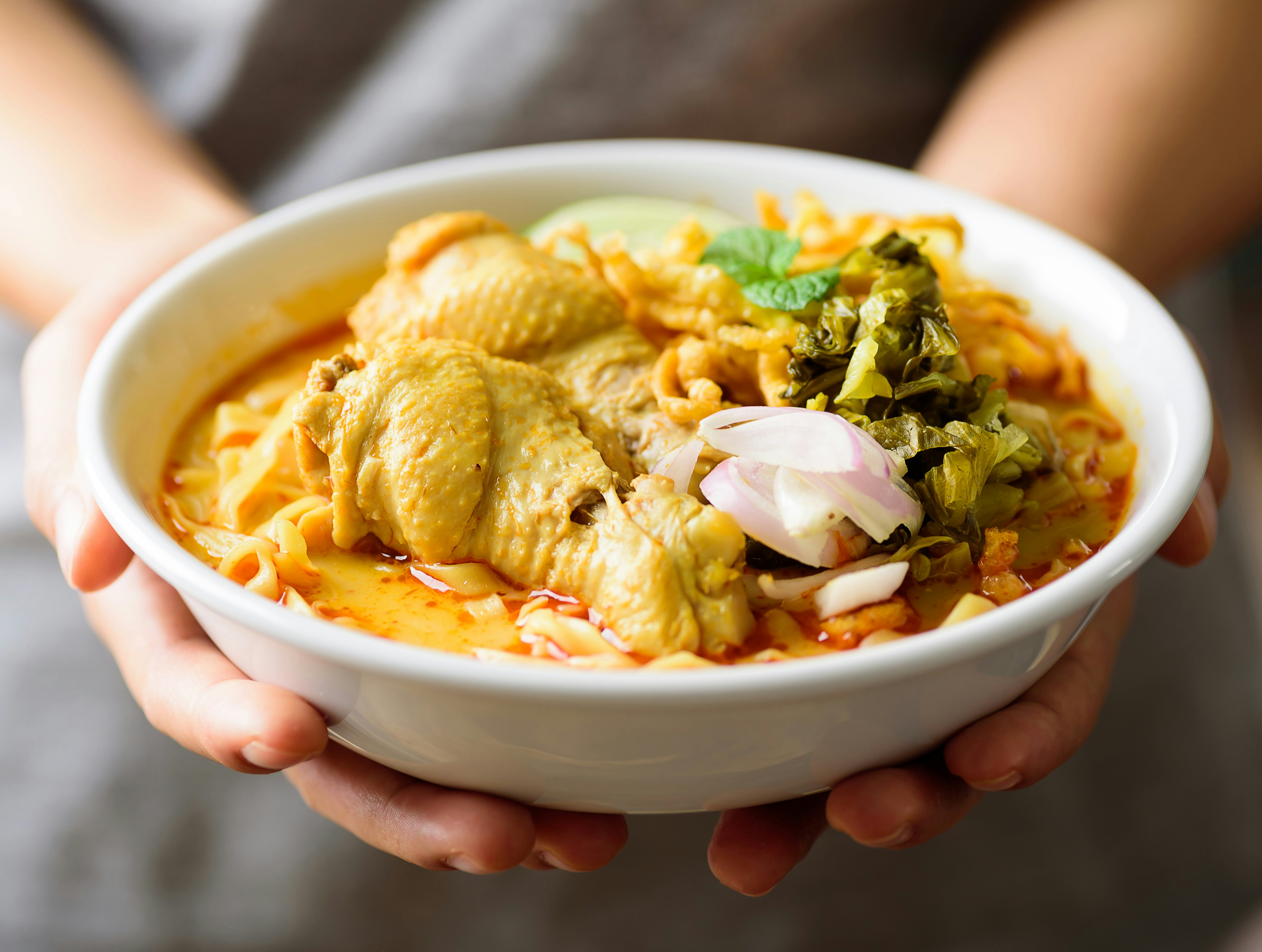 Two hands hold out a bowl of steaming khow suey, a Burmese soup dish containing noodles, vegetables and meat, in a creamy, yellow-coloured broth.