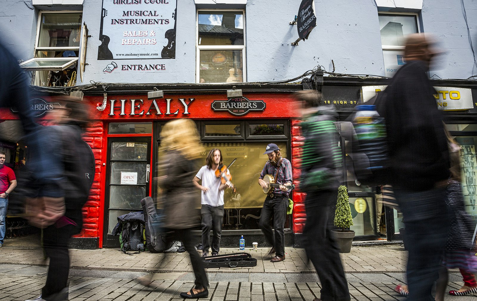 Two buskers play in front of the red facade of a barbershop in Galway. People pass in front of them, blurred by the camera. Galway, Ireland.