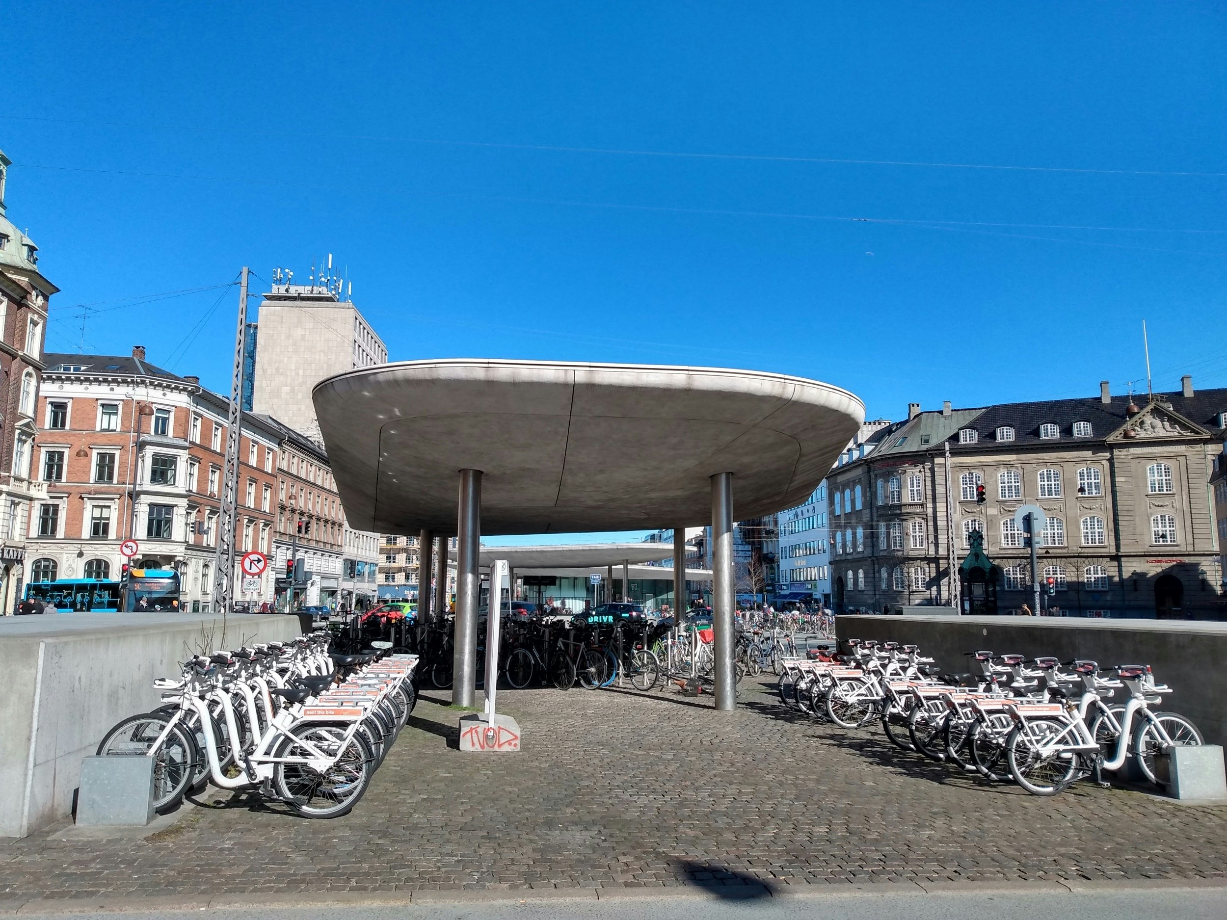 Many white bicycles are docked outside a city-centre metro station.