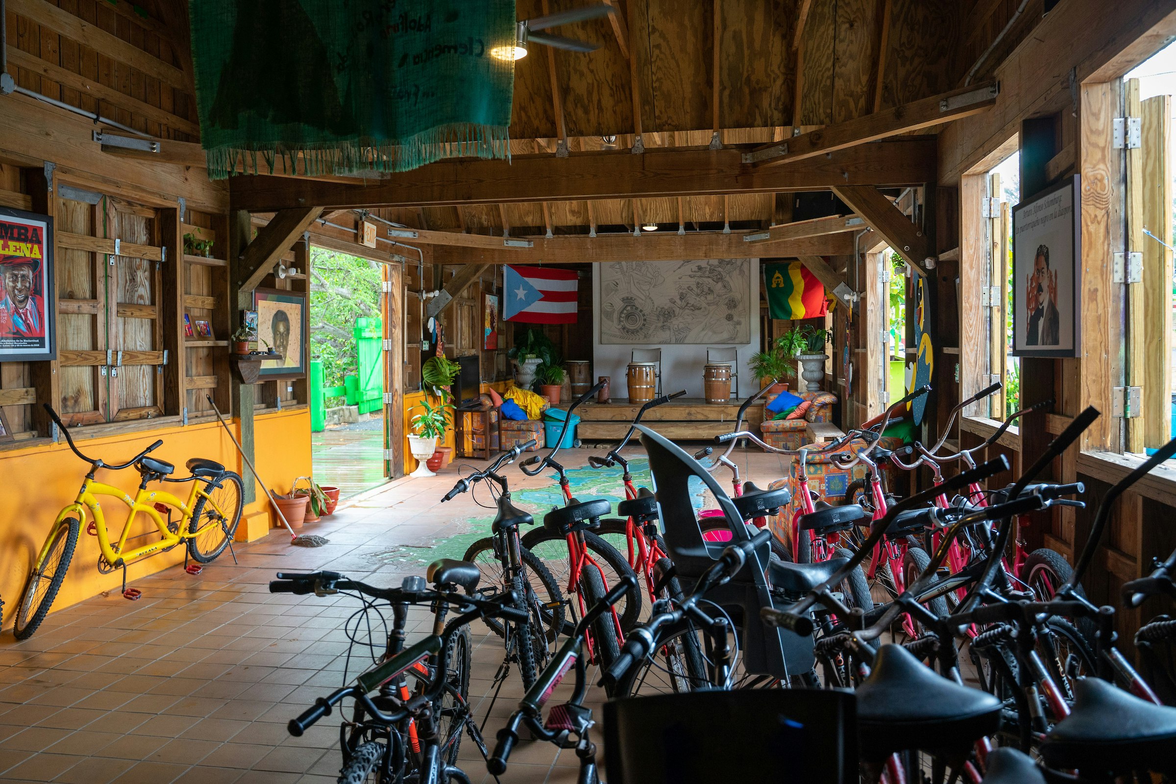 Interior of the COPI Cultural Center that houses a row of bikes towards the back of the building. The front there are two drums sitting on a stage; there is a Puerto Rico flag on the wall behind the stage; Sustainable Puerto Rico 