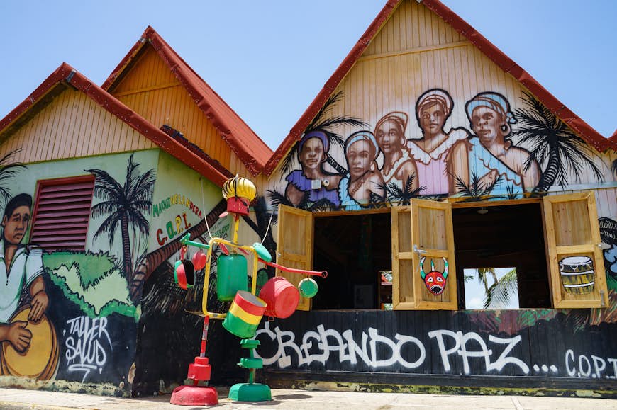 Ett par byggnader är täckta med bilder av kvinnor med huvuddukar och en man som spelar trumma;  framför byggnaden finns en färgstark struktur gjord av grytor och stekpannor;  Hållbart Puerto Rico 