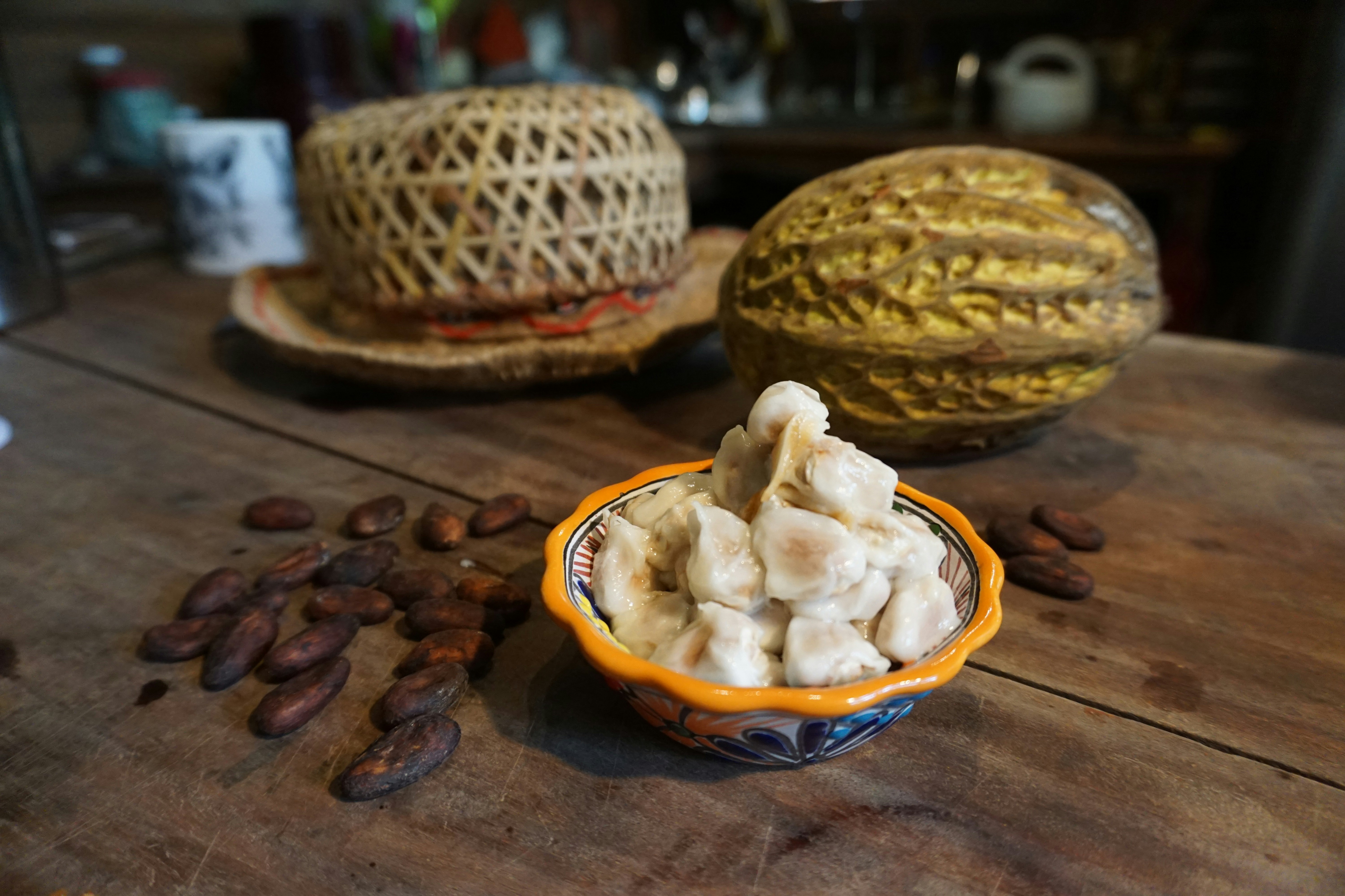 A pile of cacao fruit pods sit in a bowl, next to the bowl are roasted cacao beans. In the background is a full cacao fruit and a straw hat. 