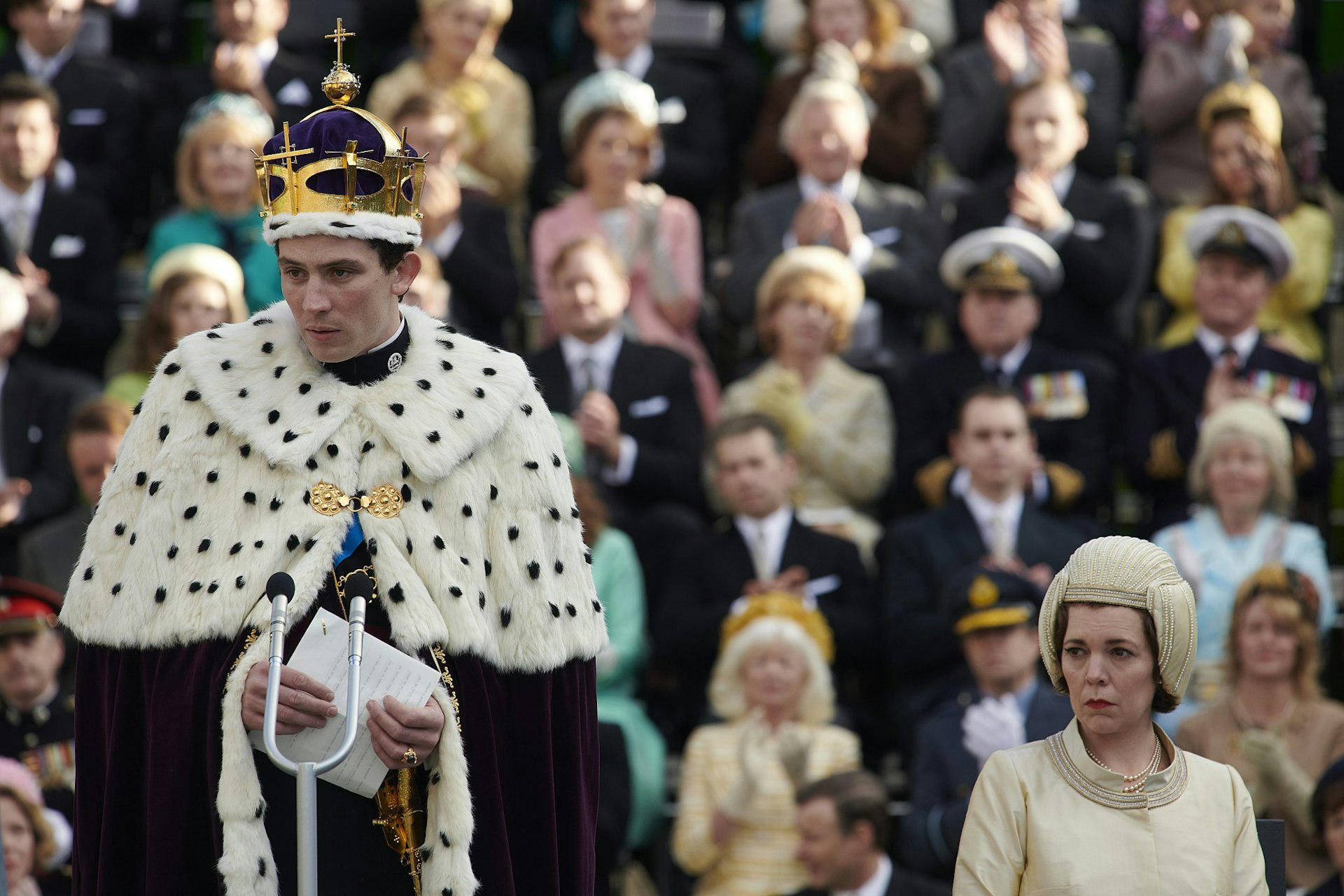 A still from The Crown season 3, depicting Prince Charles’s investiture as the Prince of Wales at Caernarfon Castle; Prince Charles is wearing ceremonial robes while the Queen sits in the background looking unhappy.
