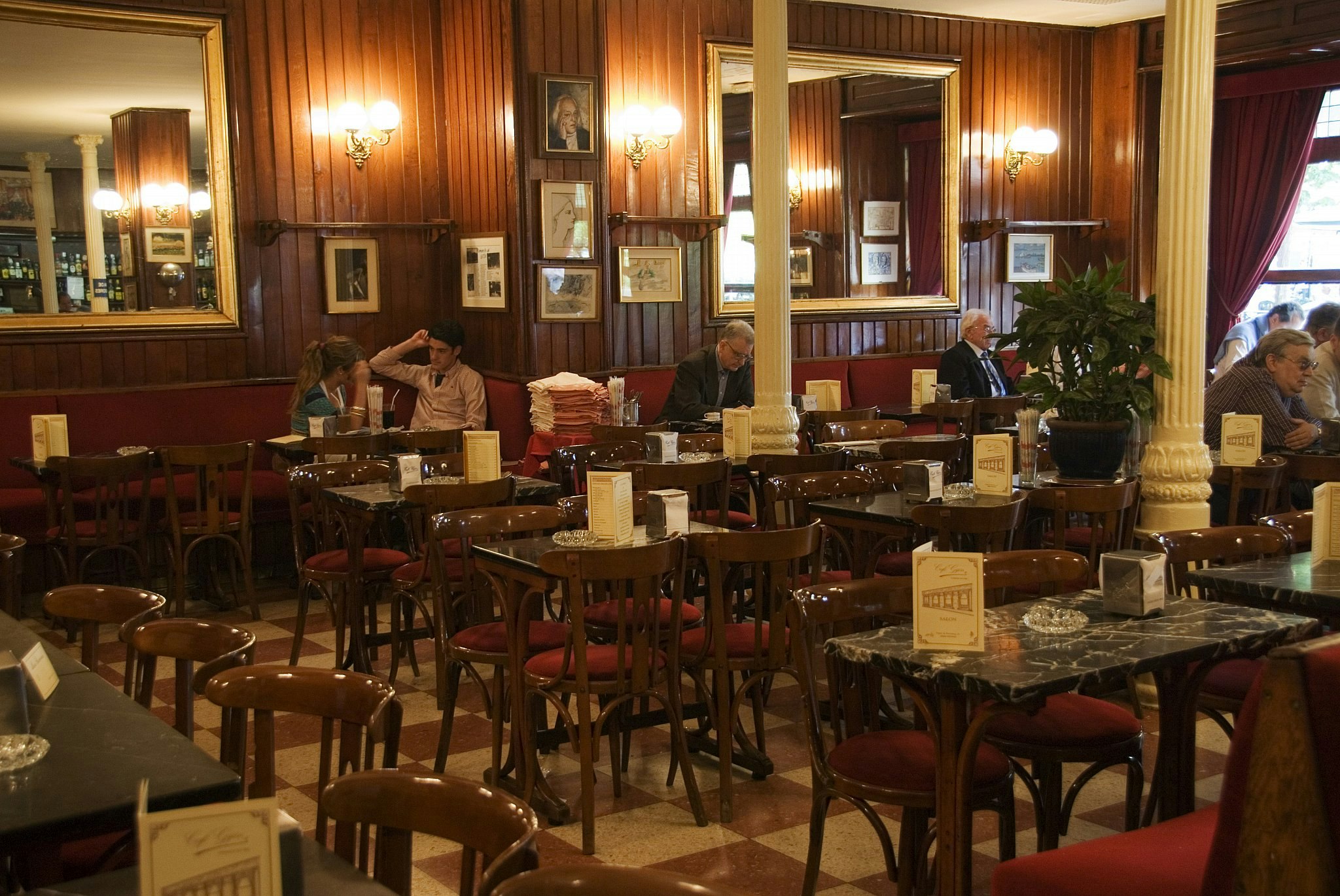People inside Café Gijón, Madrid, which has a white and red tiled floor, dark wood seats with red cushions, panelled walls and white columns.