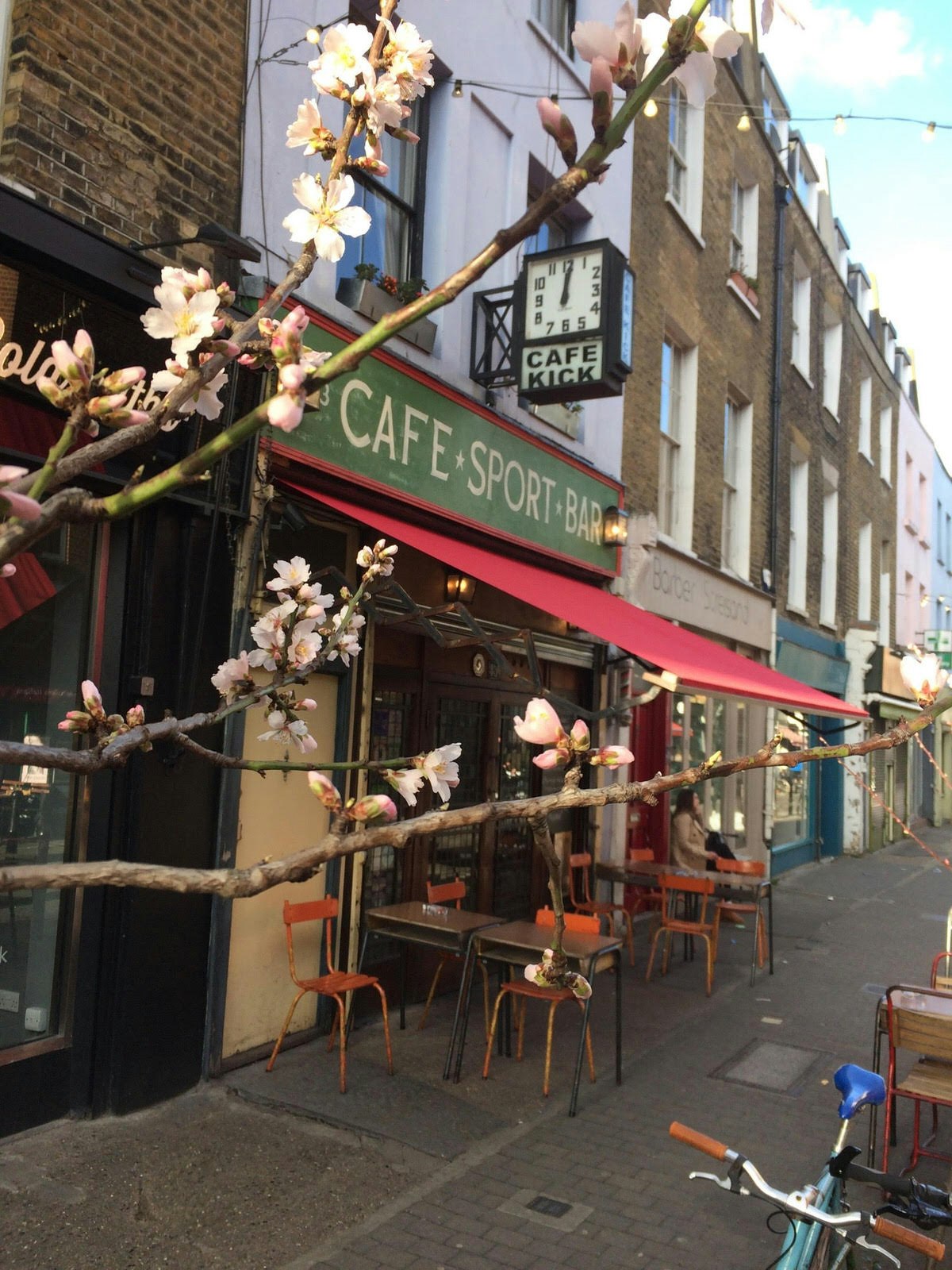 The exterior of Cafe Kick sports Bar in Exmouth Market. There is a branch of a cherry blossom in the foreground, slightly obscuring the cafe which has lots of outdoor seating