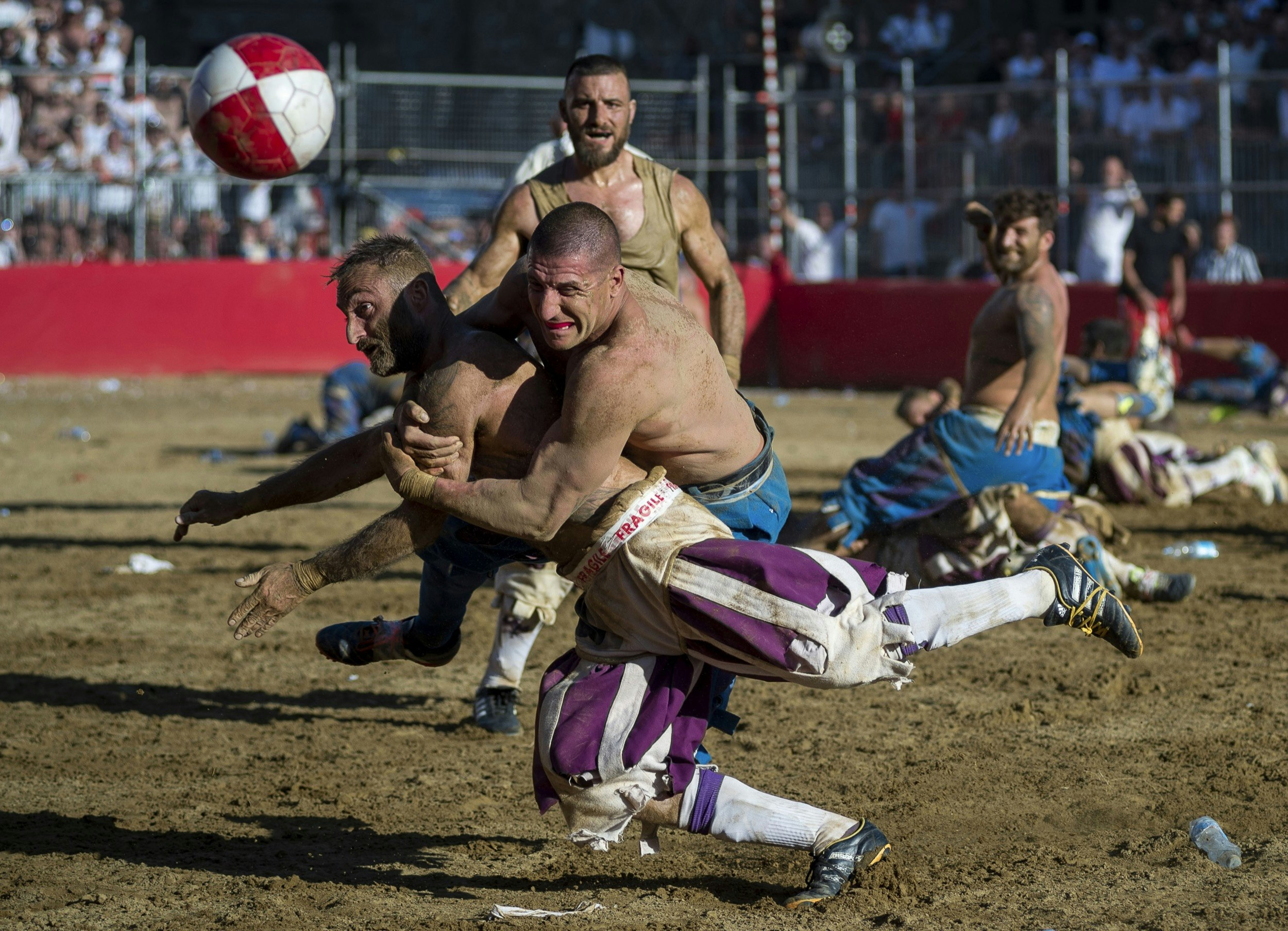 A man is tackled in a muddy arena as both men - and others around them - watch a ball bouncing erratically; unique sporting events