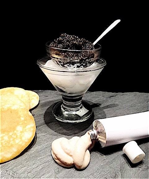 A tube of calf brain cream is photographed next to a small wine glass of caviar filled with ice. There are small crackers to the left of the caviar. 