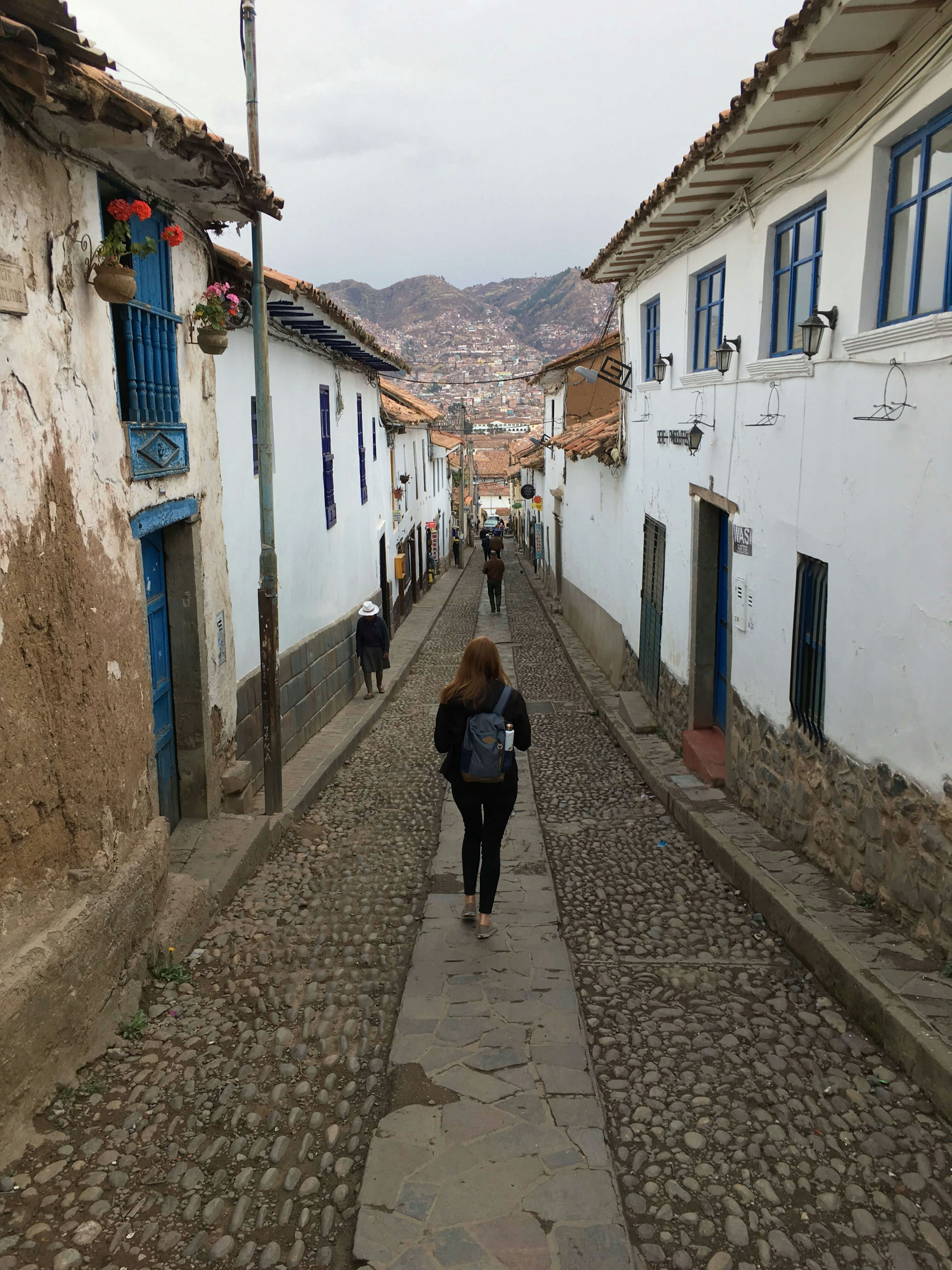 A woman walks down a cobblestone street, surrounded by white walls. The mountains are in the distance.