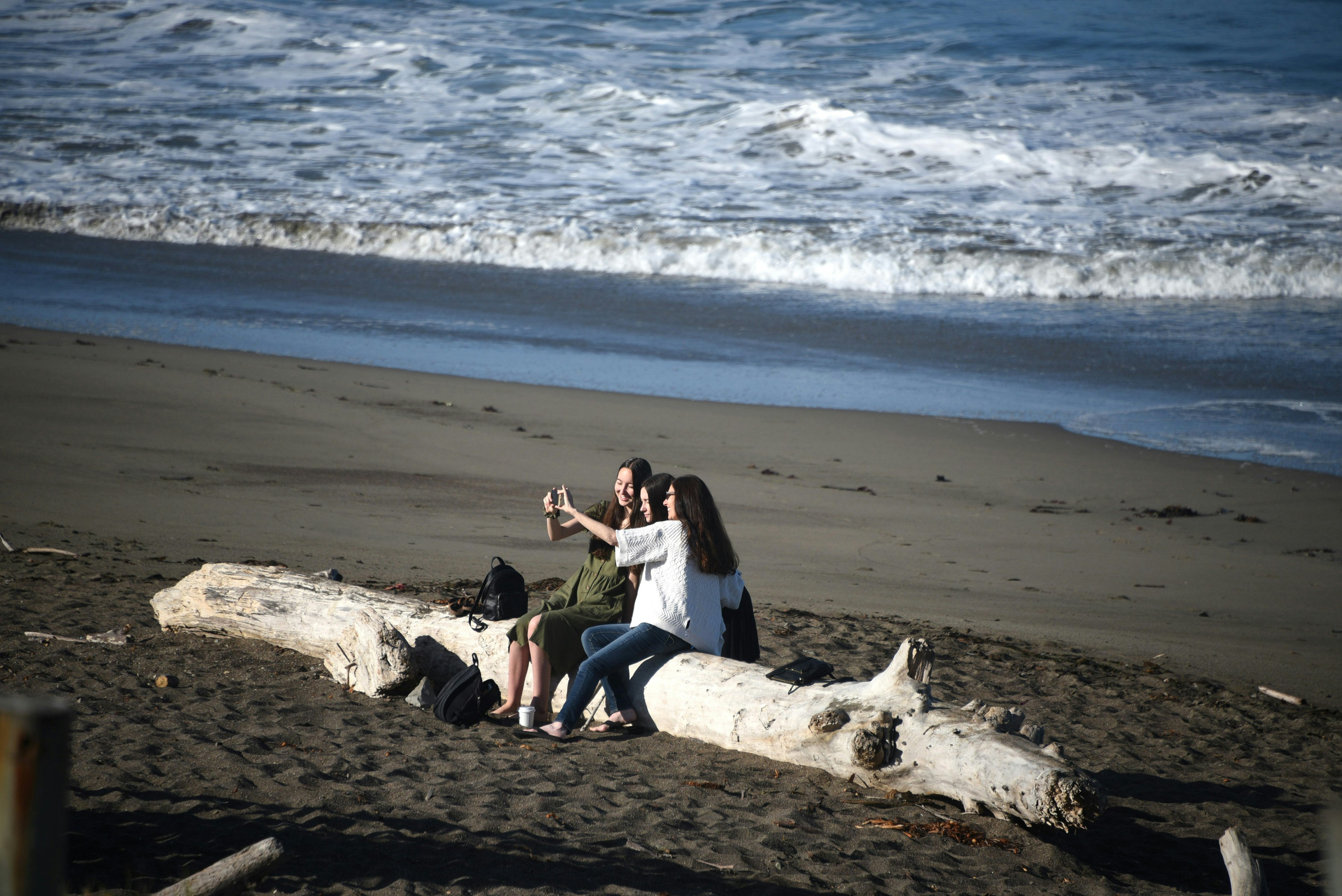 Tre vänner poserar för en selfie när de sitter på en drivvedsstock på en strand;  Kalifornien glass