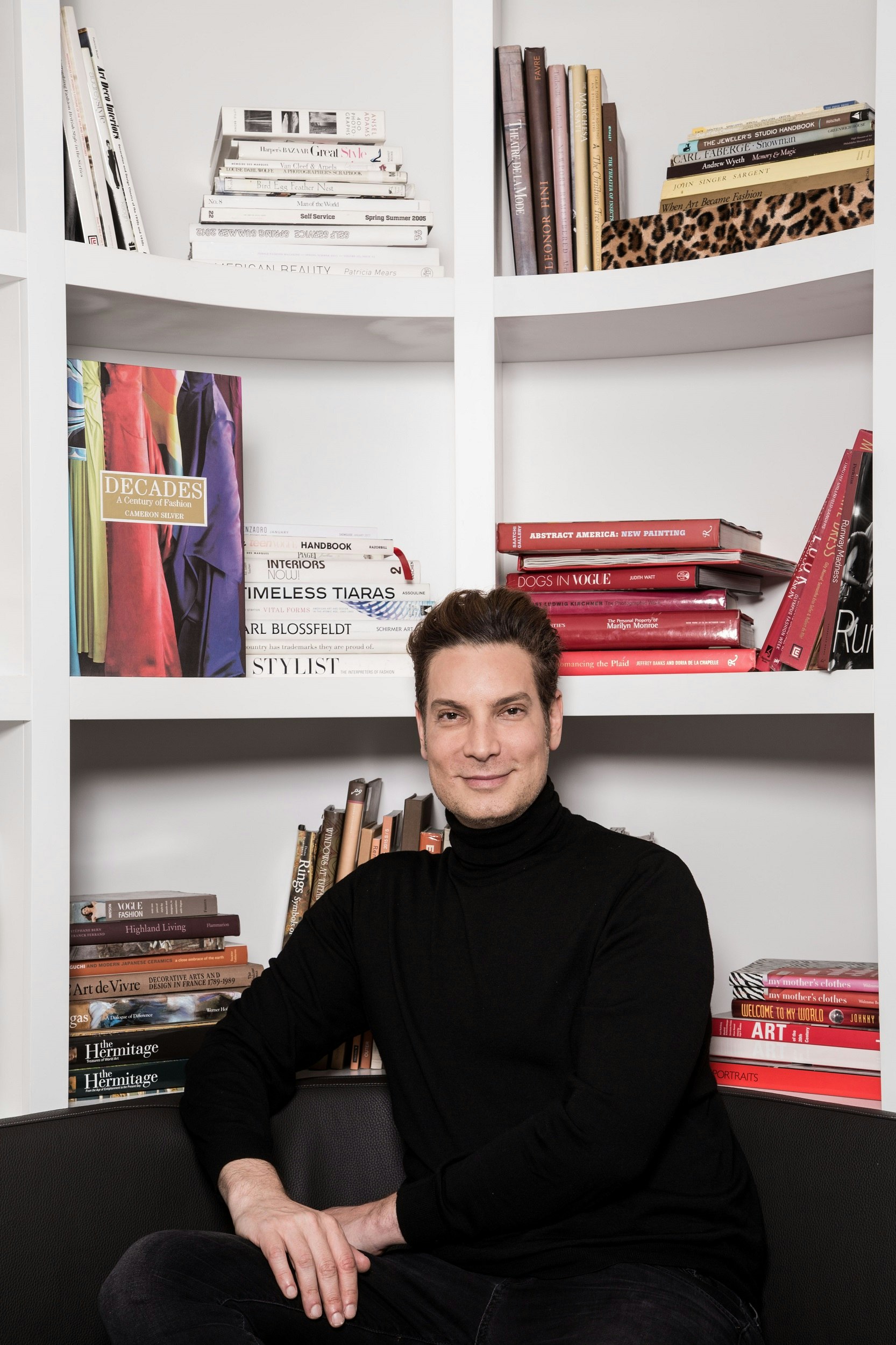 White man wearing a black round-neck top sits in front of a book shelf and smiles at the camera