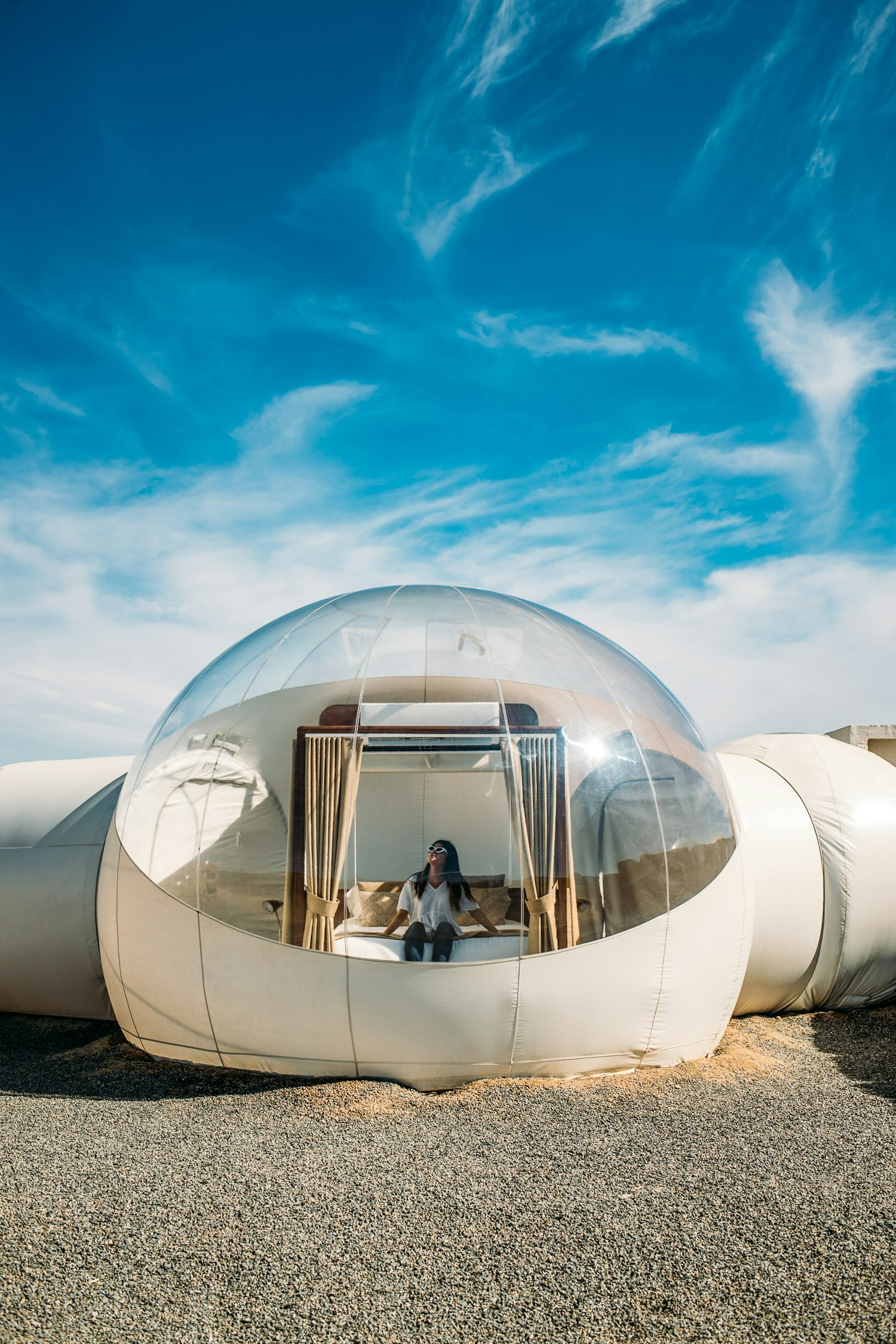 A woman peers out of the clear ceiling of a bubble tent, with blue skies above