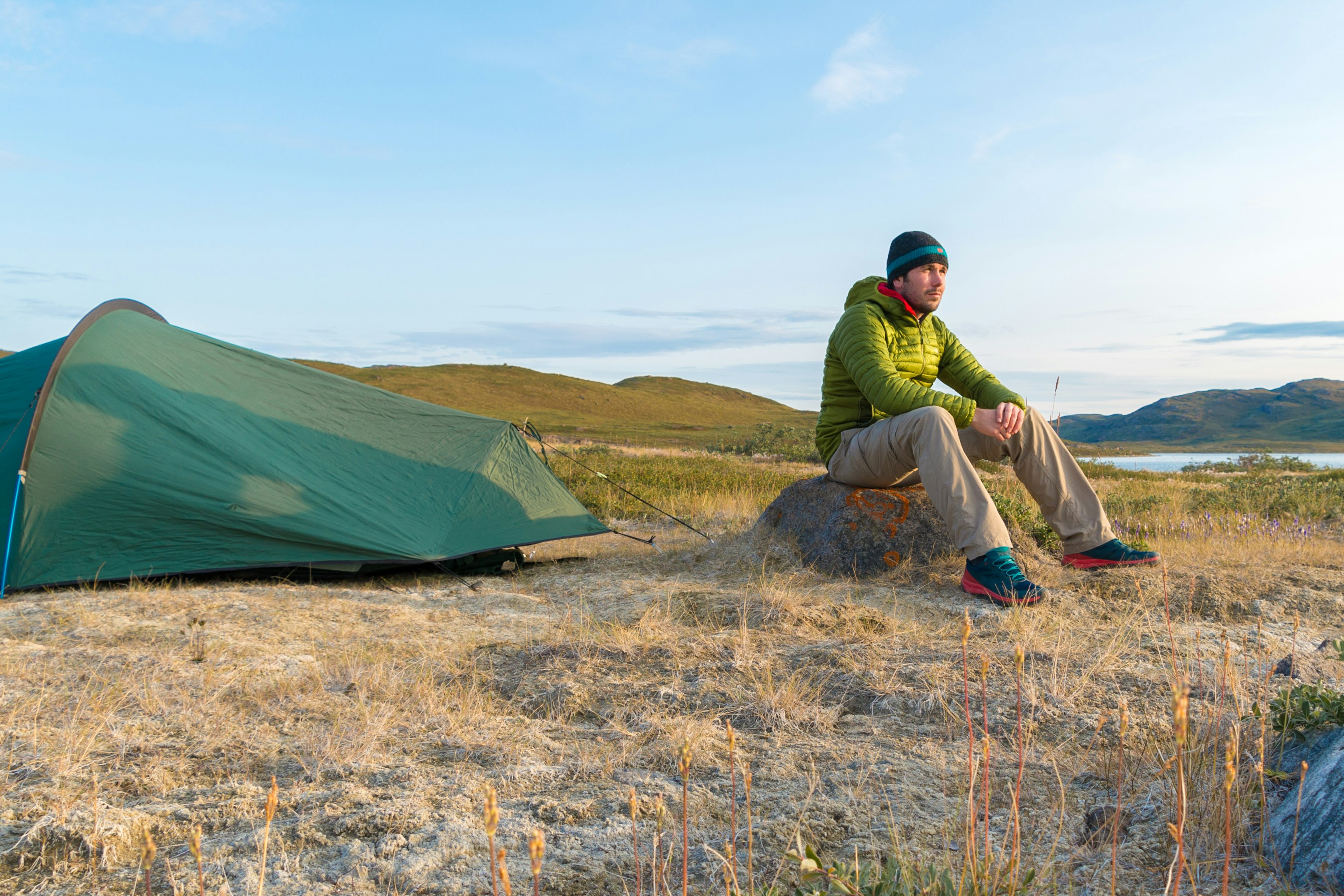 Peter sits on a rock next to his green tent admiring the Arctic scenery of lakes and green hills.