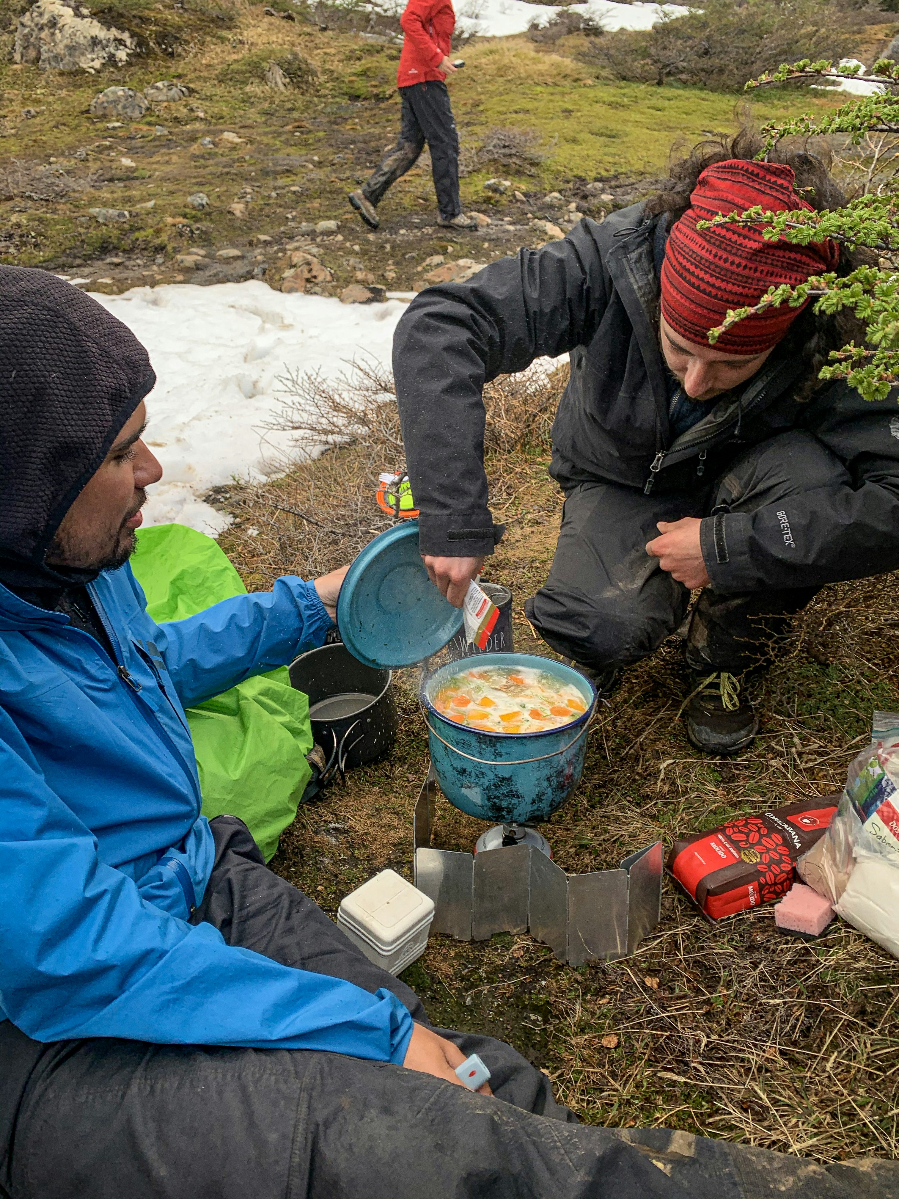 A campsite scene; two members of the trekking team are cooking a stew in a large saucepan on a camping stove.