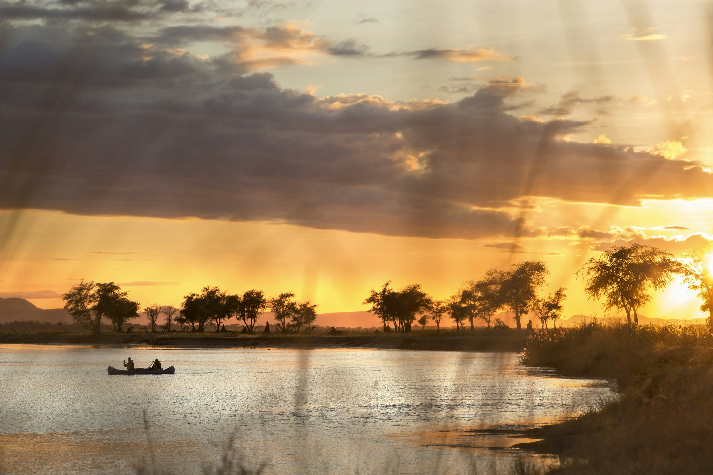 A canoe sits in a curve of the Zambezi River, with acacia trees silhouetted along its shore; the sky is golden, with dark clouds cutting across it.