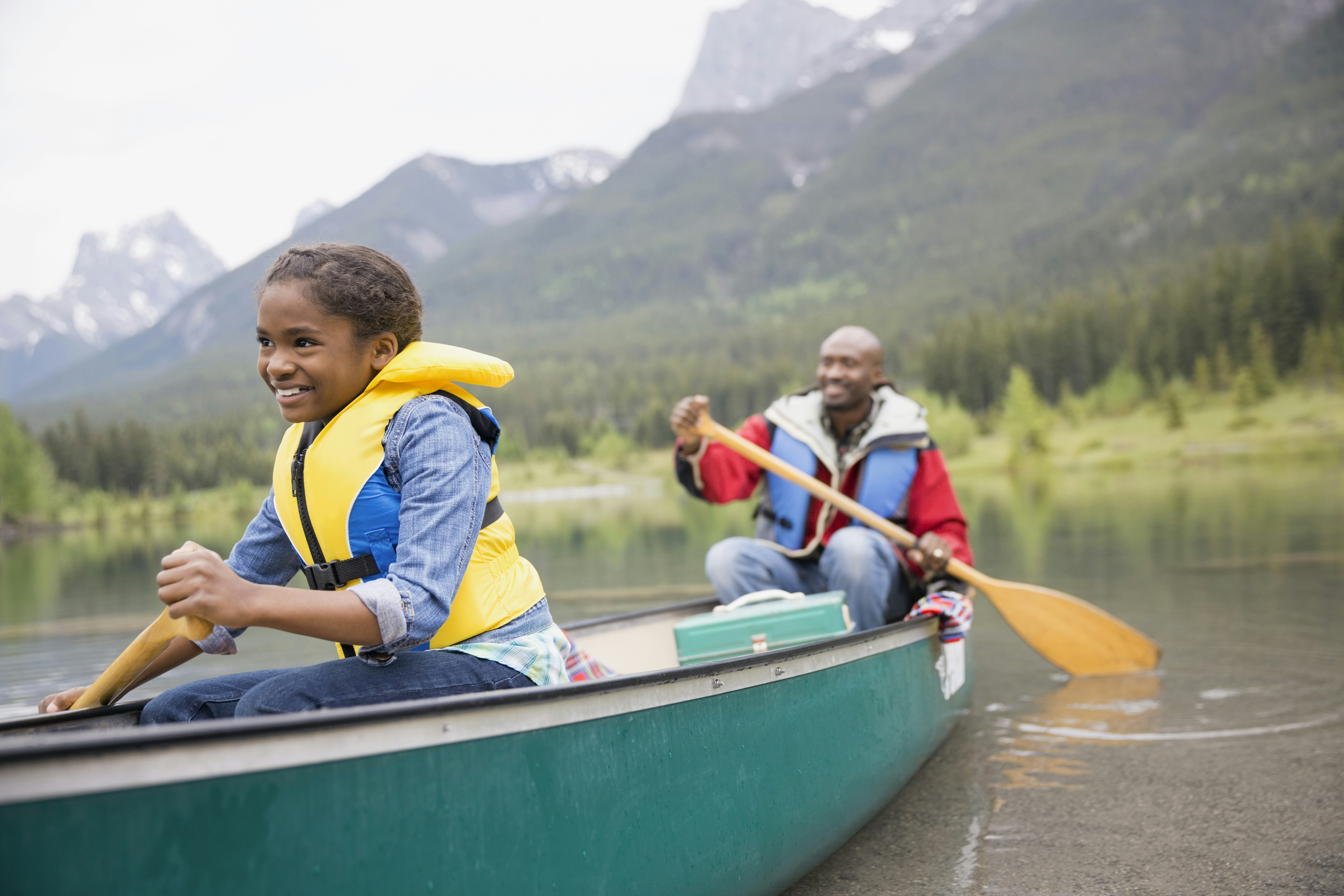 A man and a girl paddle a canoe through a lake with mountains rising in the background; kids outdoor adventures