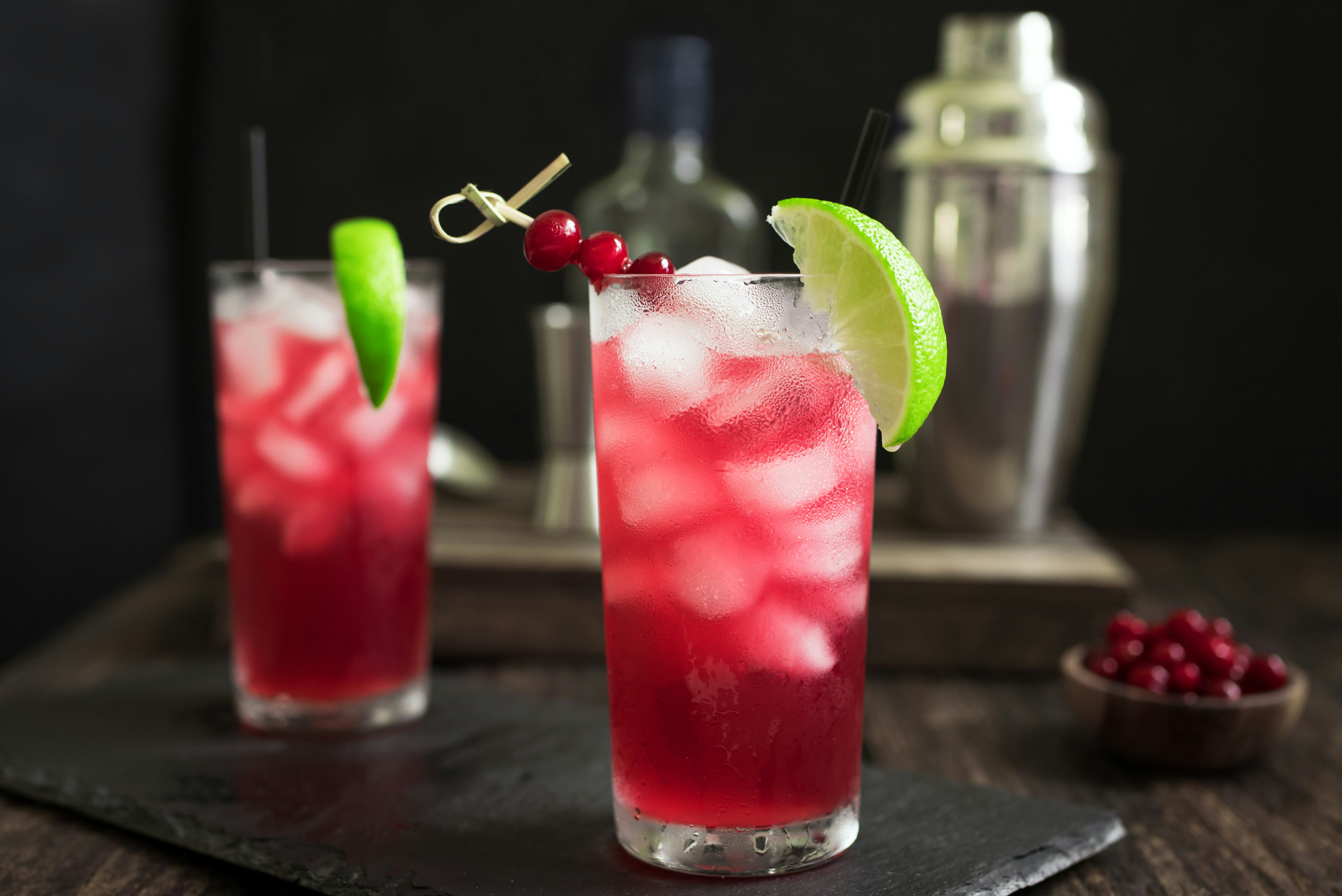 Ett par höga glas fyllda med tranbärsjuice och vodka och is.  Det finns en limeklyfta på glasets kant, ett långt svart sugrör och en rad tranbär.