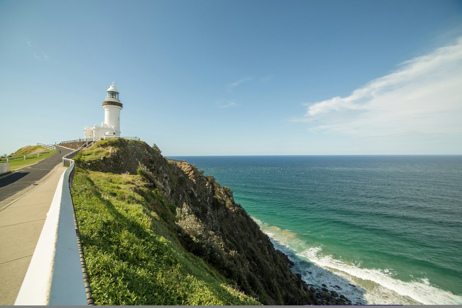 A road leads towards a white lighthouse at the end of the Cape Byron Walking Track; it stands on a rocky cliff, with waves crashing beneath.