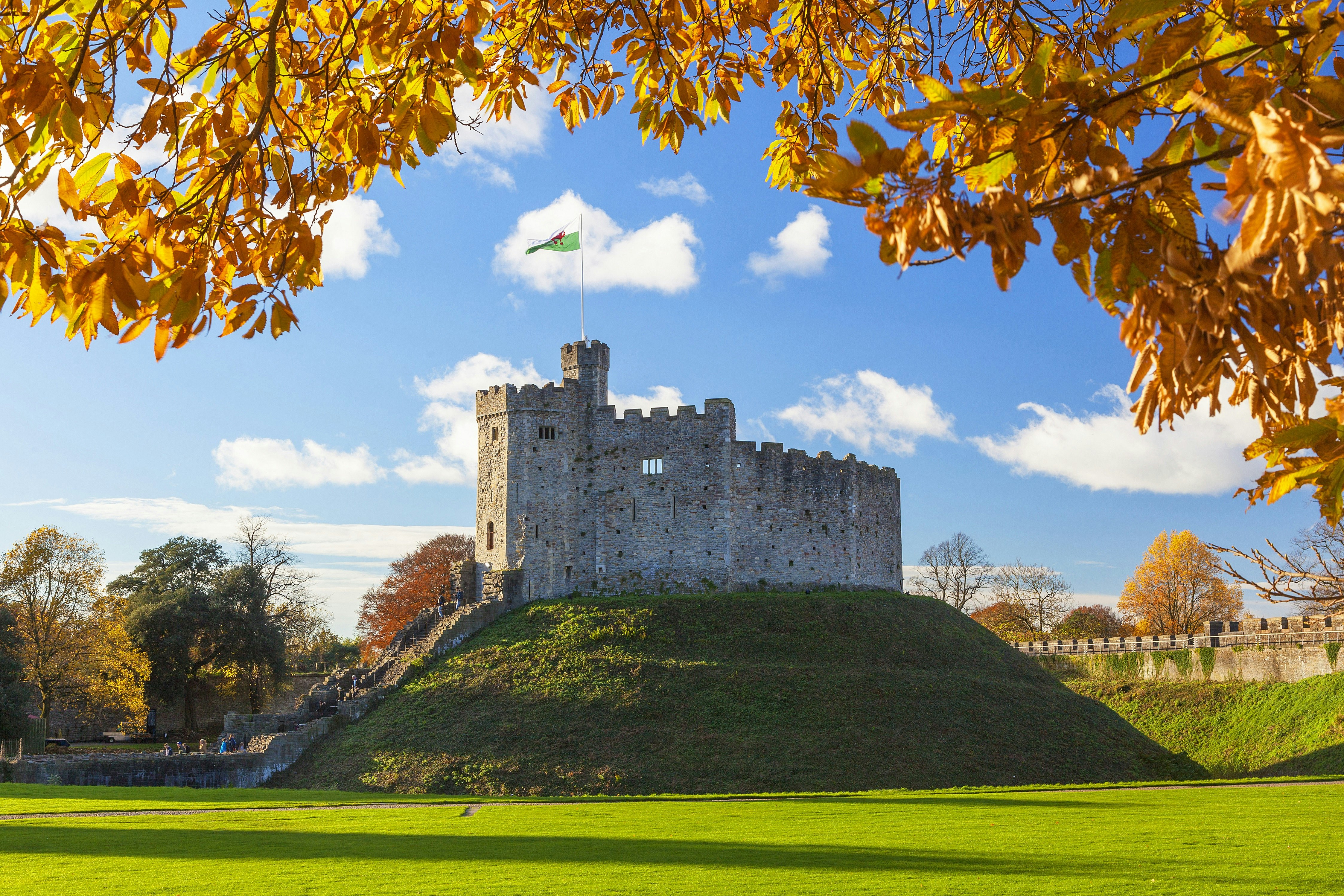 A view of Cardiff Castle, a stone castle atop a small hill, with the top of the image framed by autumn leaves. The sun shines in the background.