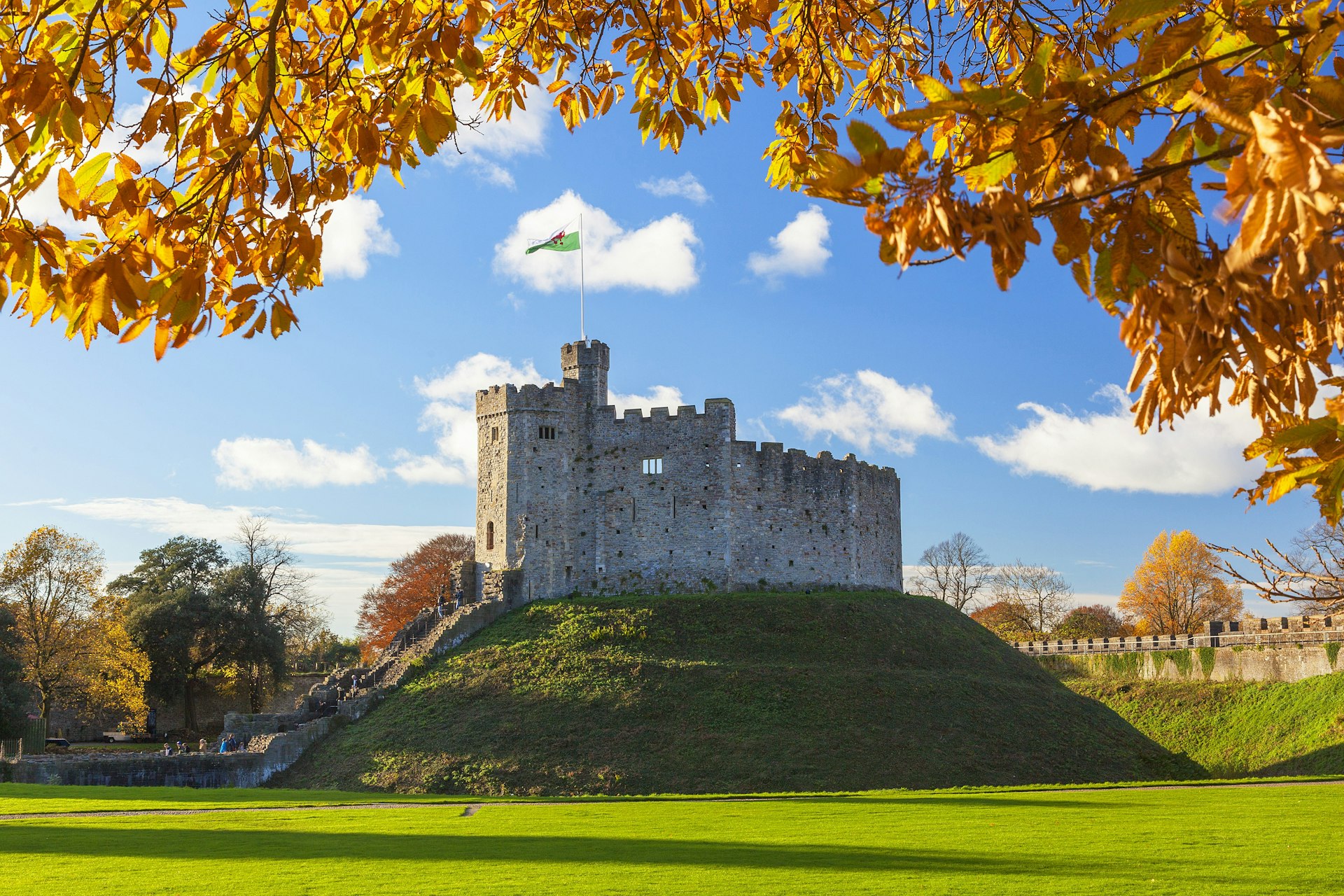 A view of Cardiff Castle, a stone castle atop a small hill, with the top of the image framed by autumn leaves. The sun shines in the background.