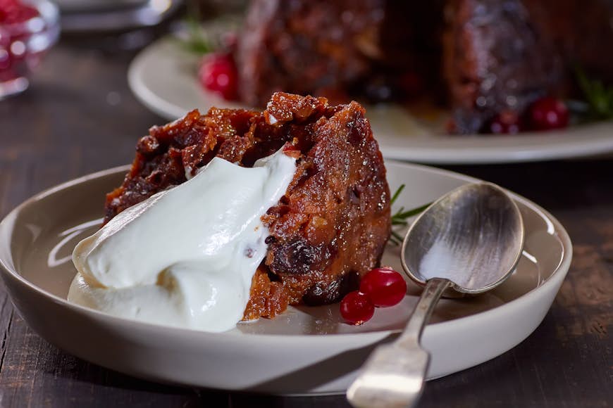 A slice of cake filled with dried fruits sit on a plate with a dollop of whip cream on top