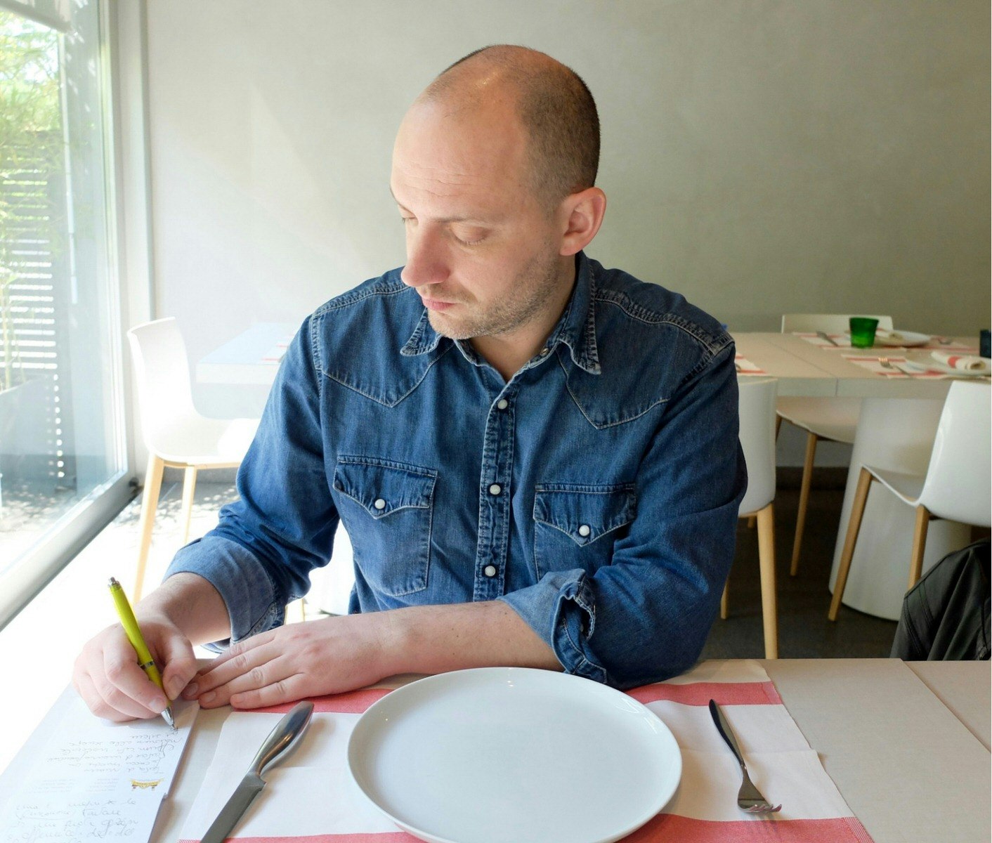 A man sitting at a table that is set for a meal. He is writing on, and looking down at, a notepad to his right