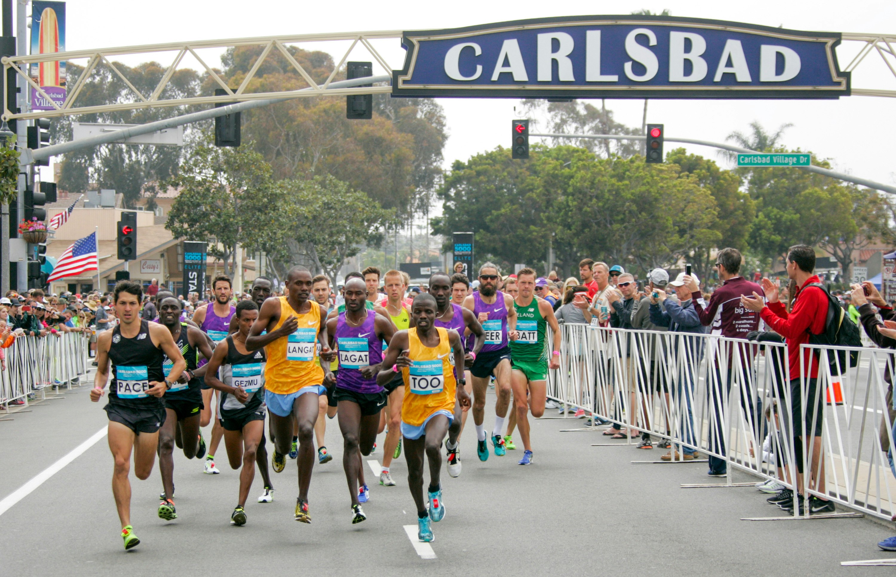 A group of male runners run past an overhead sign that says 'Carlsbad' on a closed off street in Carlsbad, California