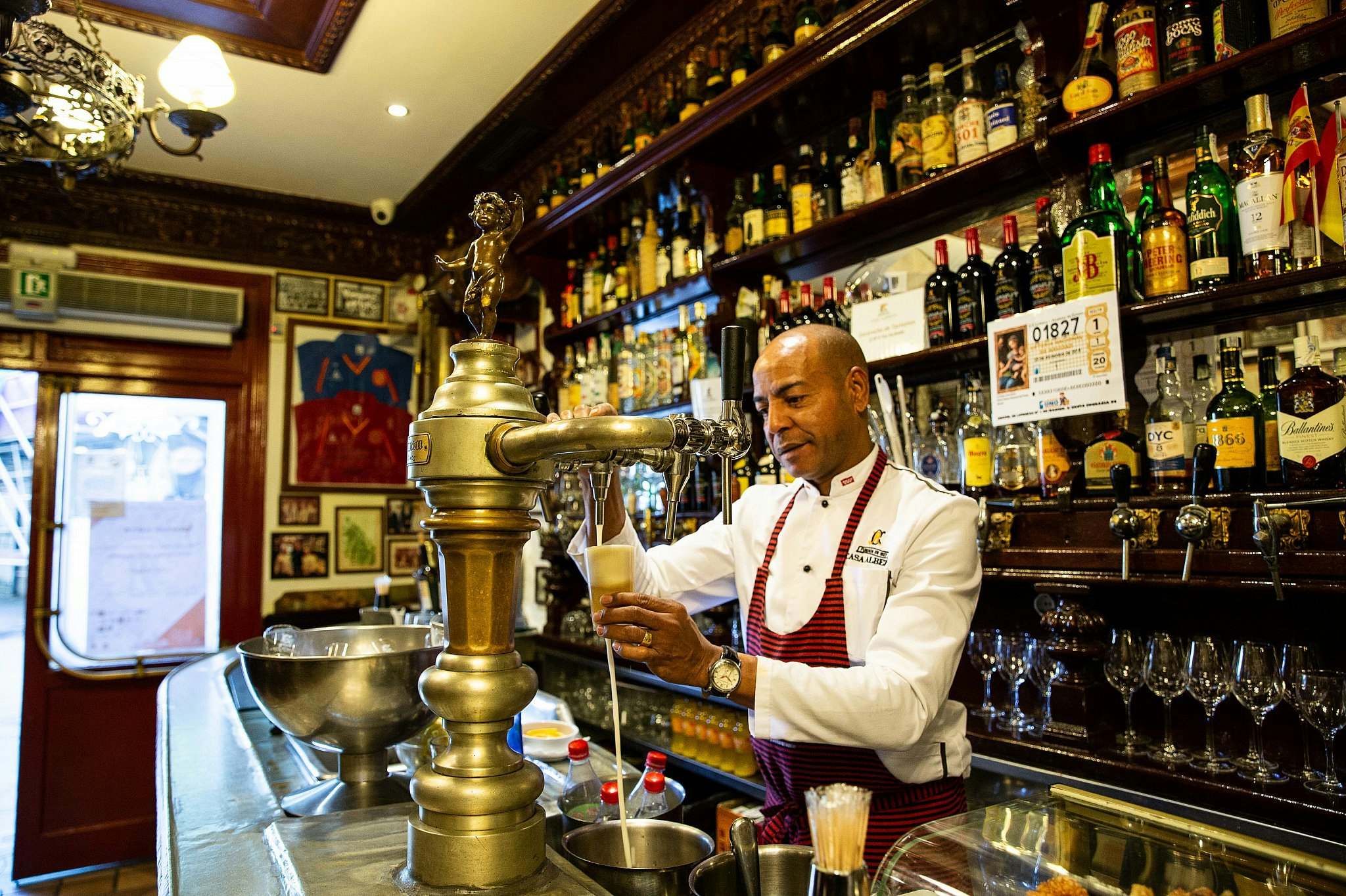 An aproned waiter pours beer from a vintage tap at Casa Alberto in Madrid; behind him, dark wood shelves are lined with bottles.