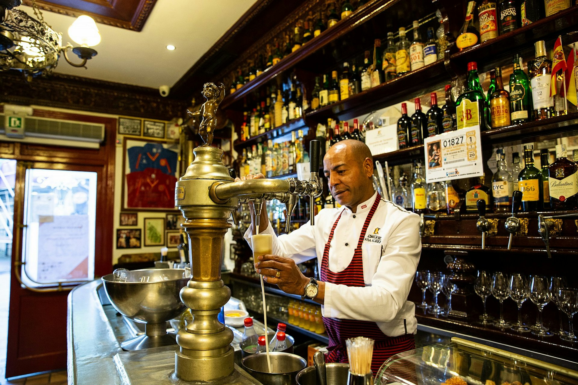 An aproned waiter pours beer from a vintage tap at Casa Alberto in Madrid; behind him, dark wood shelves are lined with bottles.