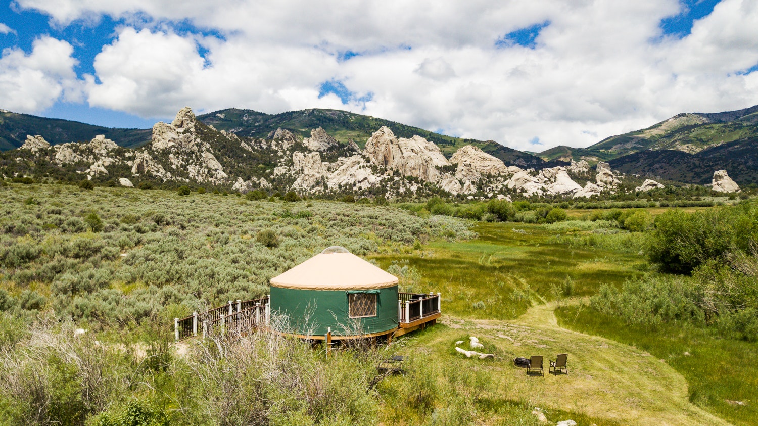 A yurt surrounded by grassland, with mountains in the background and puffy clouds above.