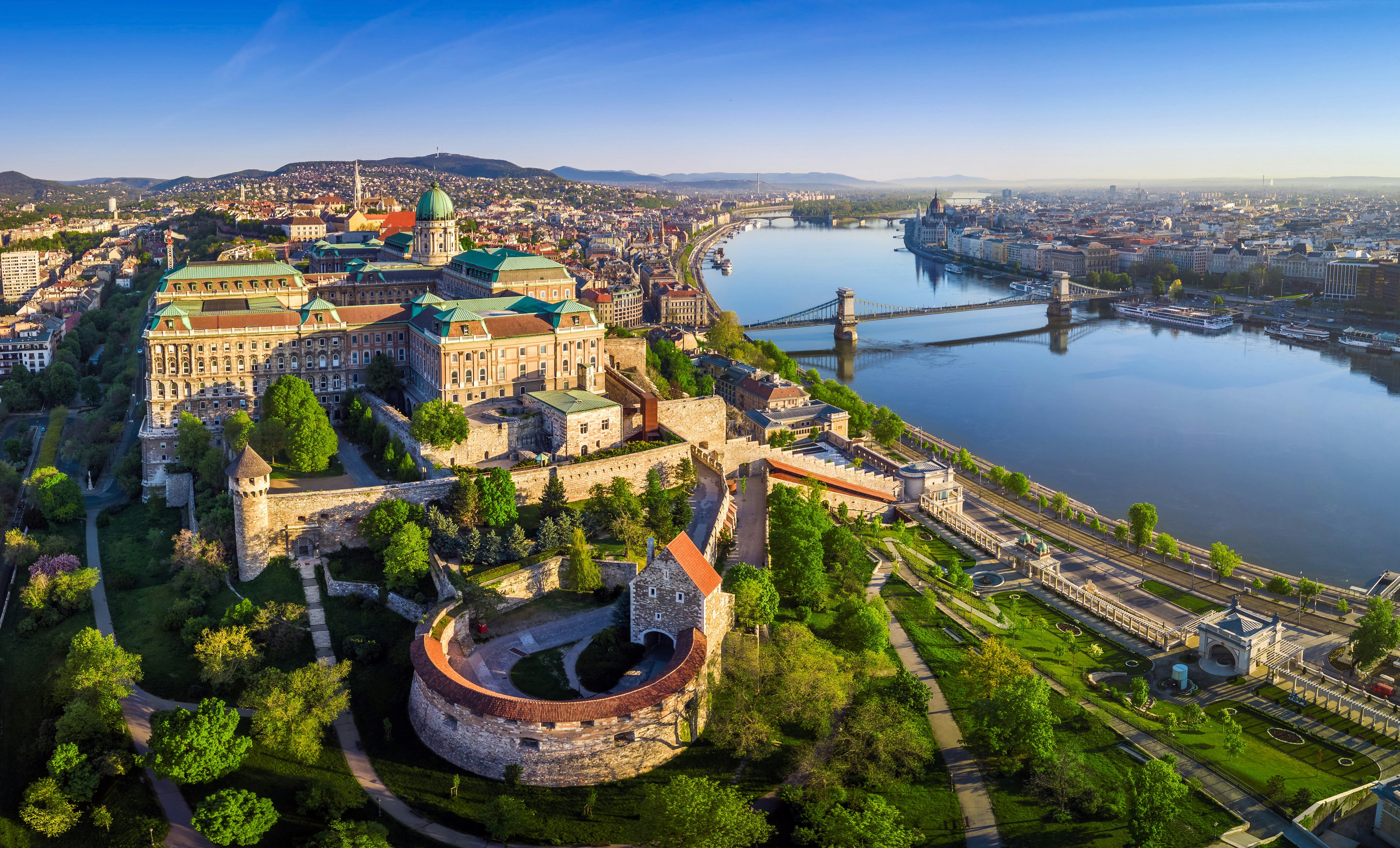 En flygfoto över Budapests slottsdistrikt en solig dag.  De gamla stenbyggnaderna är omgivna av fickor av grönska.