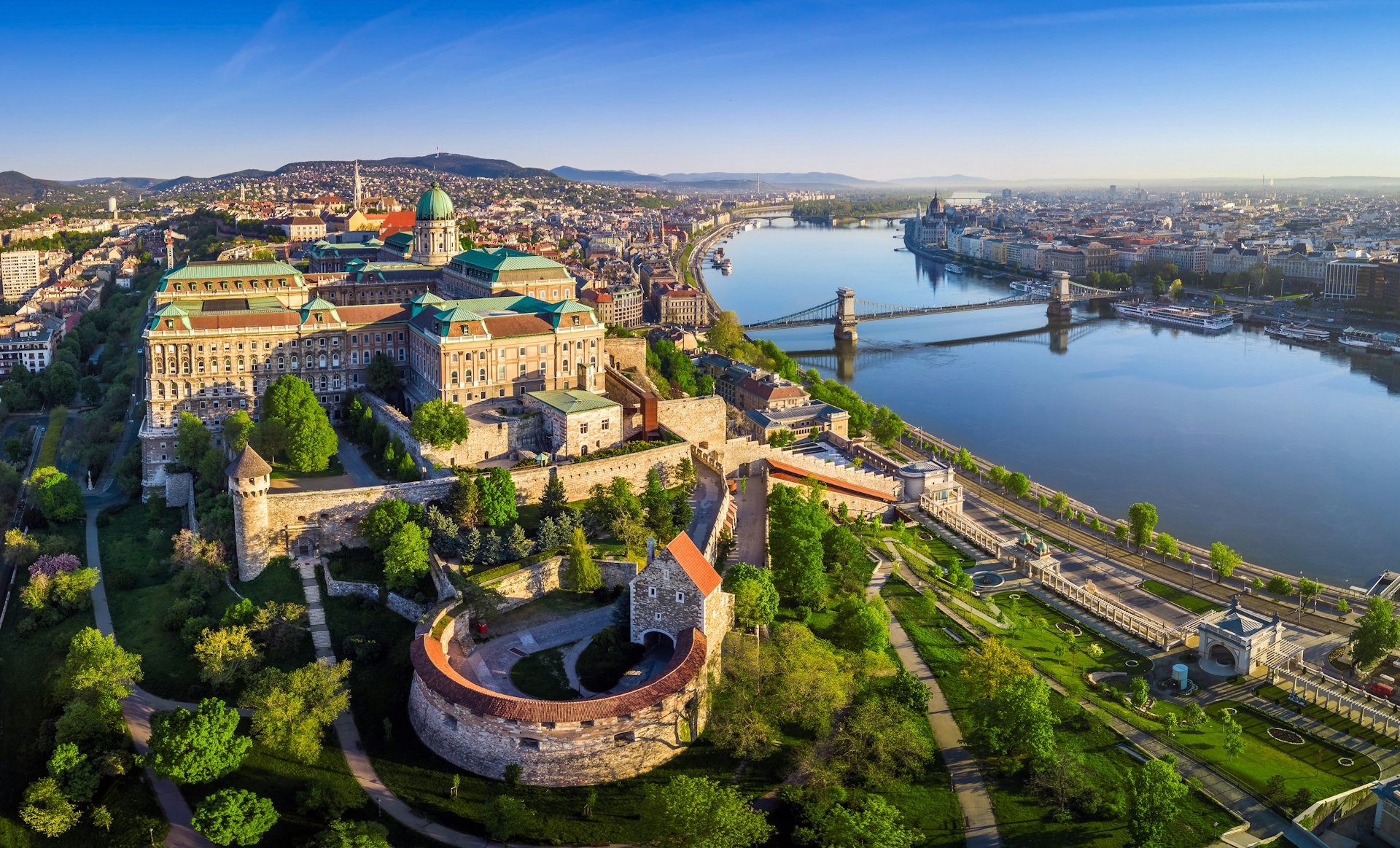 An aerial view of Budapest's Castle District on a sunny day. The old stone buildings are surrounded by pockets of greenery.