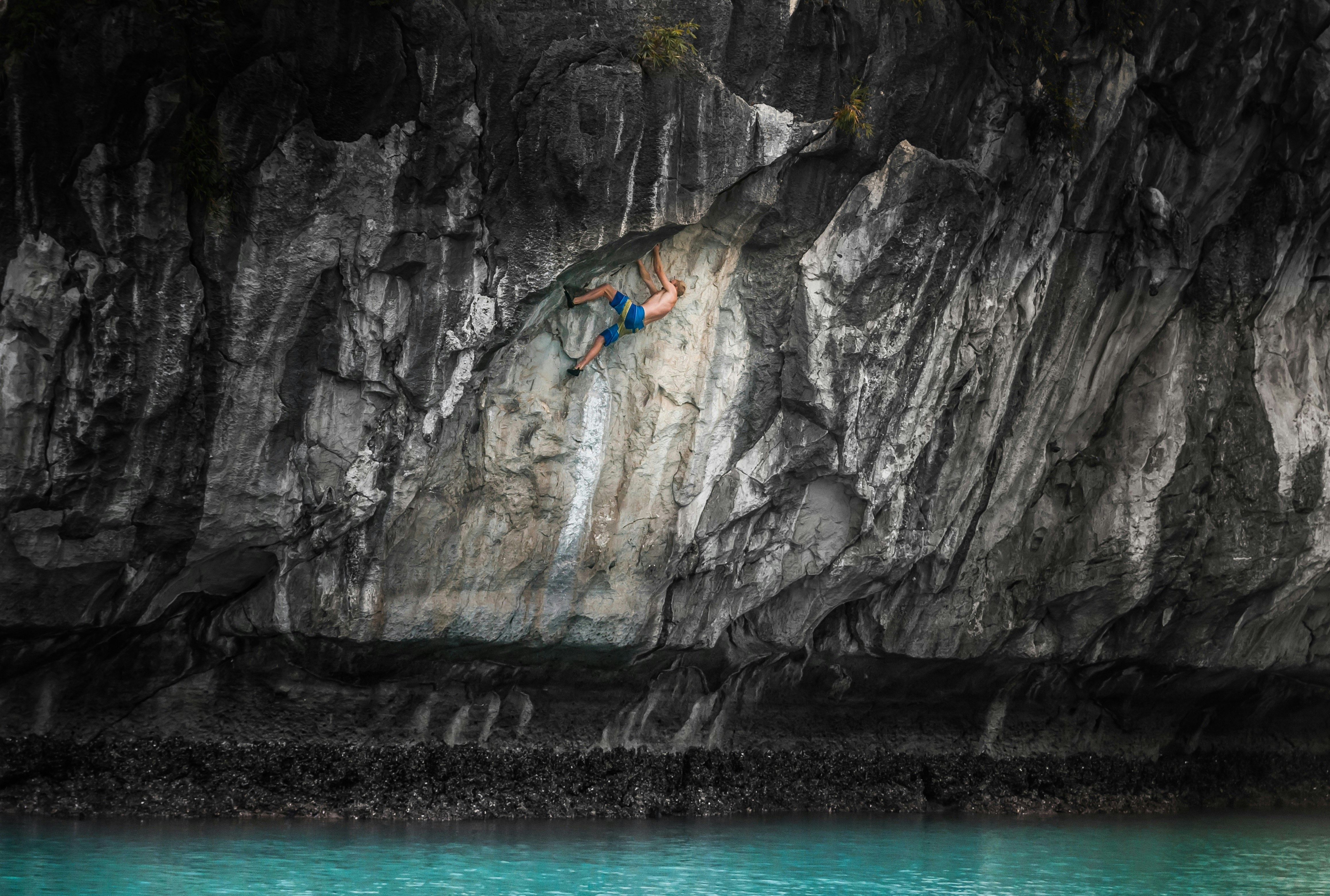 A man clambers up a sheer rock face above a turquoise sea in Cat Ba Island, Vietnam.