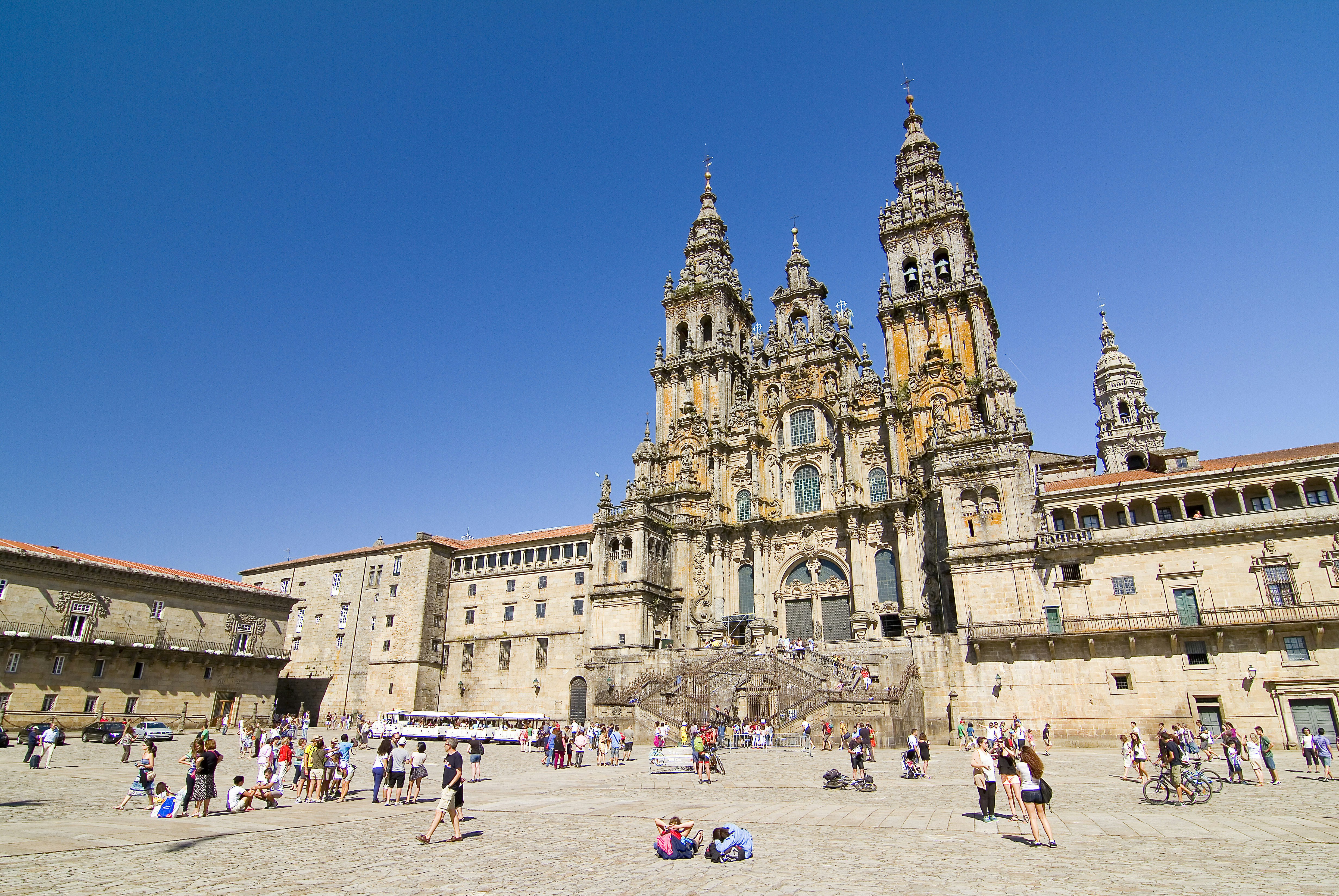A large public square is dominated by the Catedral de Santiago de Compostela; there's a bright blue sky and many people are milling around in the square.