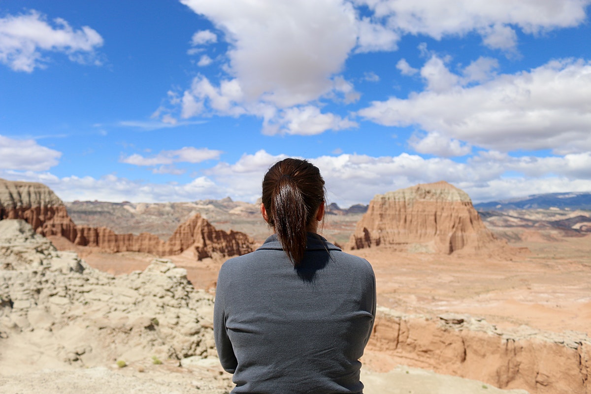 A brunette white woman sits with her back to the camera in front of the dramatic orange landforms and dragon-back ridges of Cathedral Valley