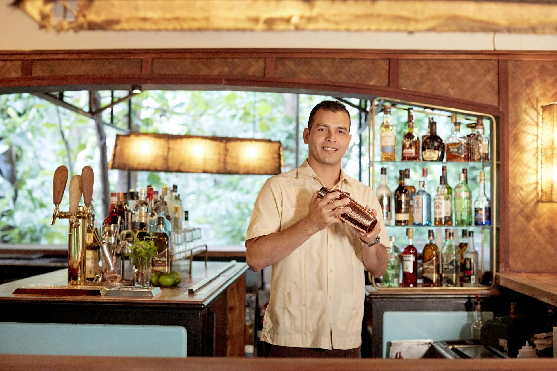 A man holding a cocktail shaker stands behind a well-stocked bar and smiles towards the camera