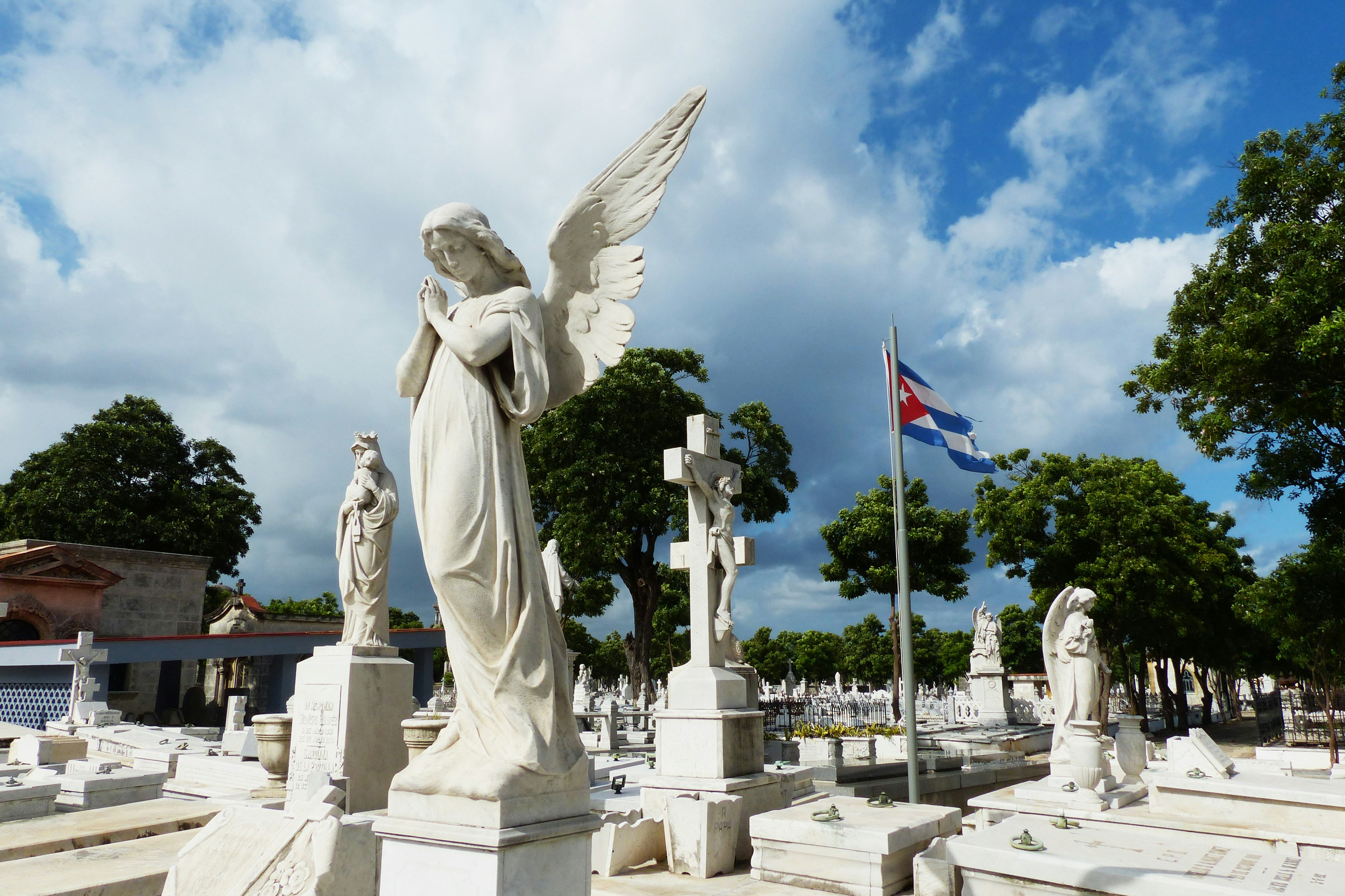 An angel carved out of stone with clasped hands and looking down sits atop a gravesite in a Cementerio Colon in Havana; Historic Havana sites