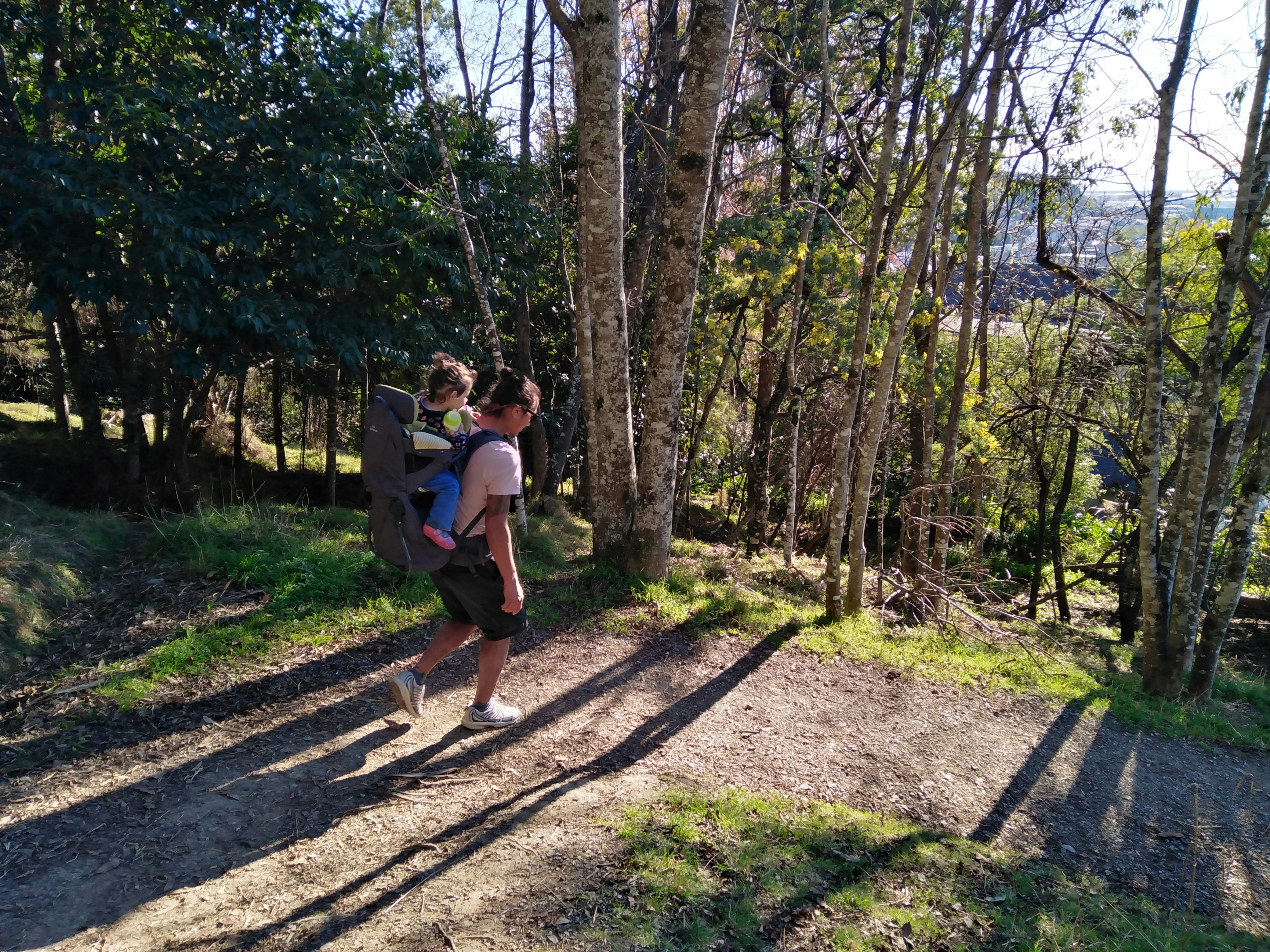 A man with a child on his back in walking down a hill in The Centre of New Zealand. The pair are walking through the woods, and their shadows are long on the ground. 