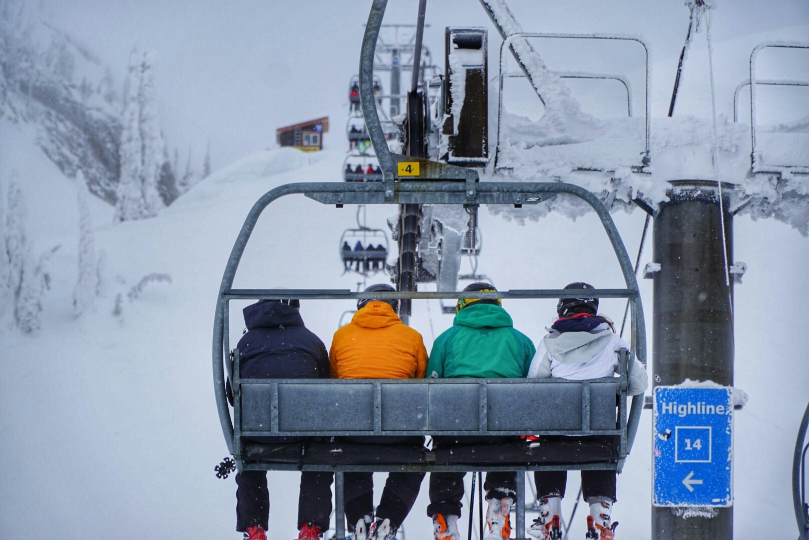 Four people in ski jackets of different colors are on a chair lift, ascending a mountain. Their backs are to the camera and it's snowing lightly.
