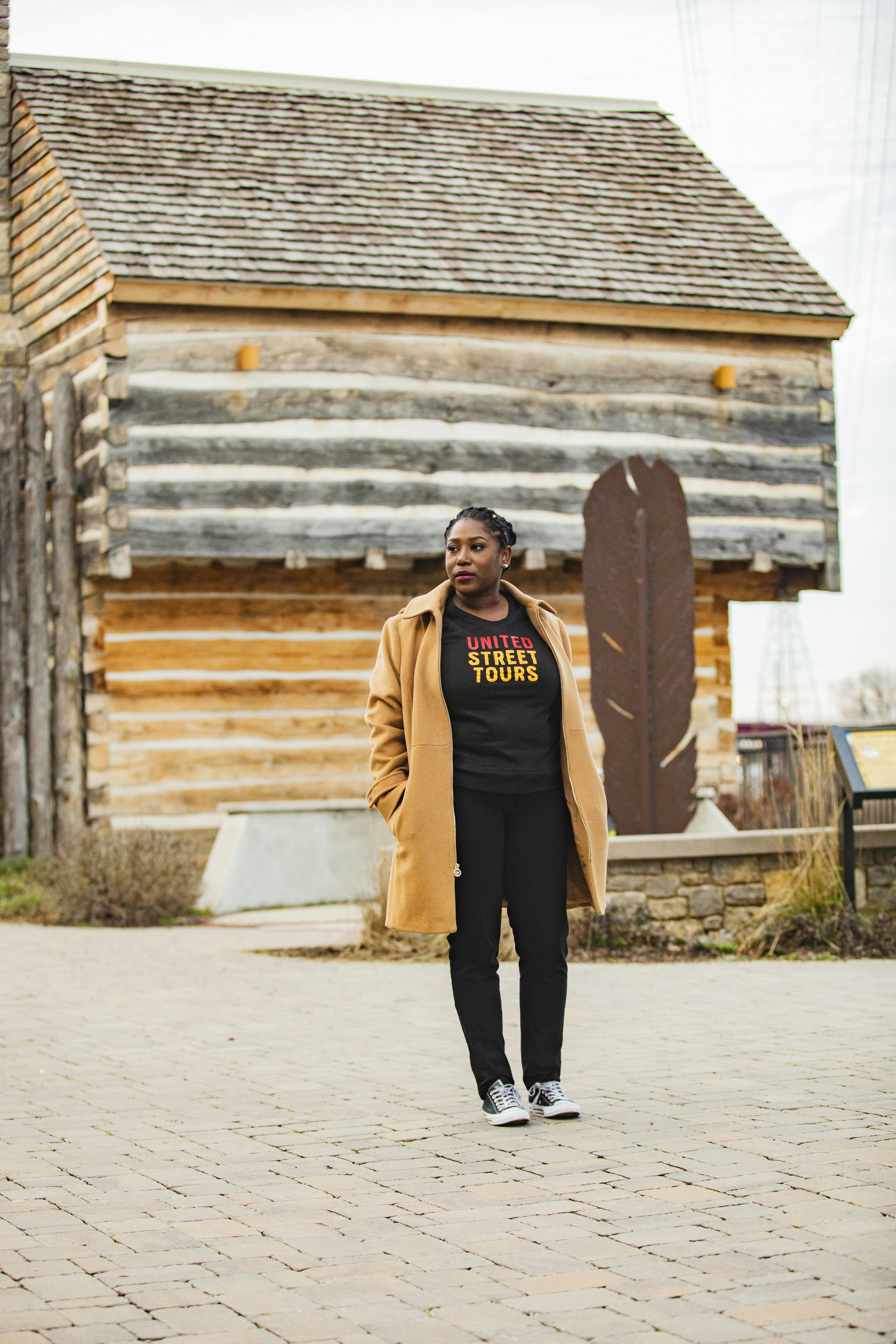 A woman stands in front of a wooden cabin wearing a camel trench coat and a black shirt that says "United Street Tours"