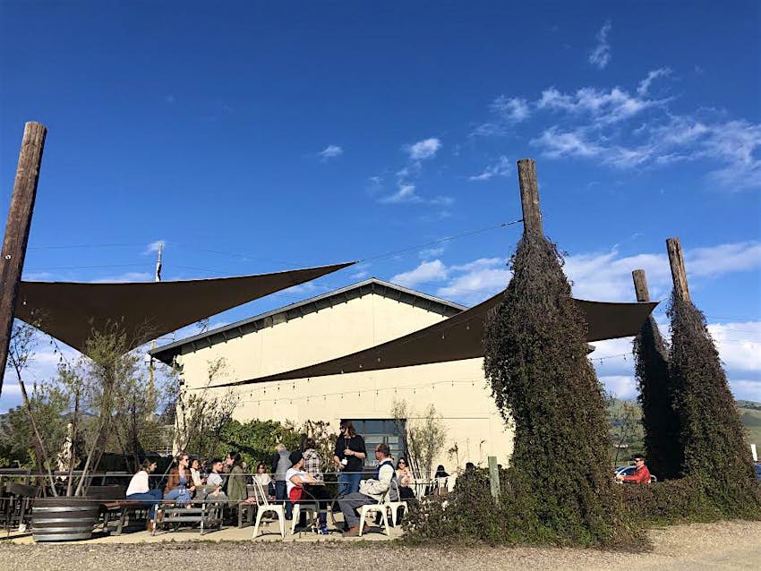 Wine tasters sit under black sun protective sails on the patio at Chamisal Vineyards in San Luis Obispo County, California by the white exterior of the tasting room itself