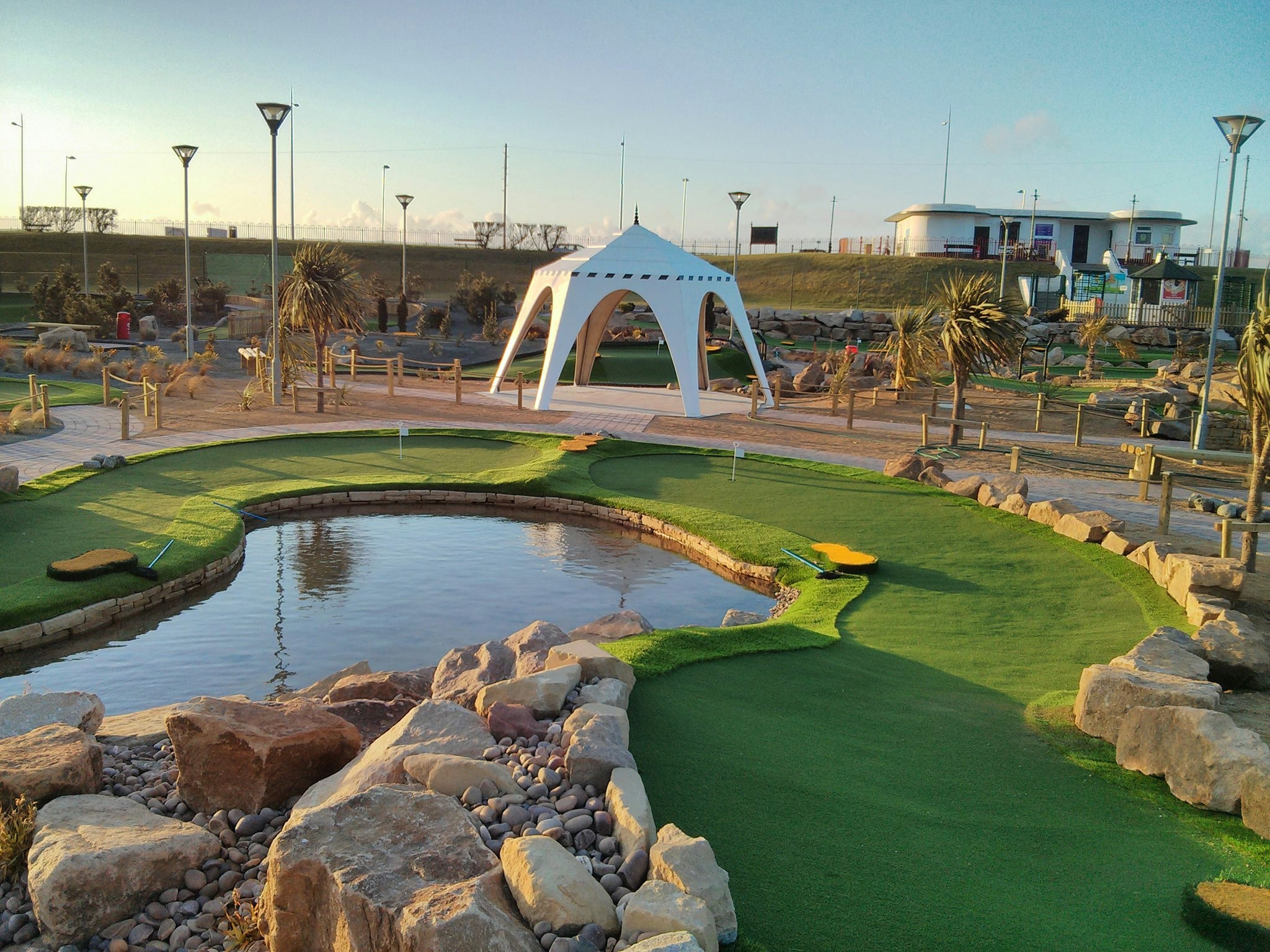 A body of water is in the middle of a green at Championship Adventure Golf, in England. An awning is visible at the center of the image.