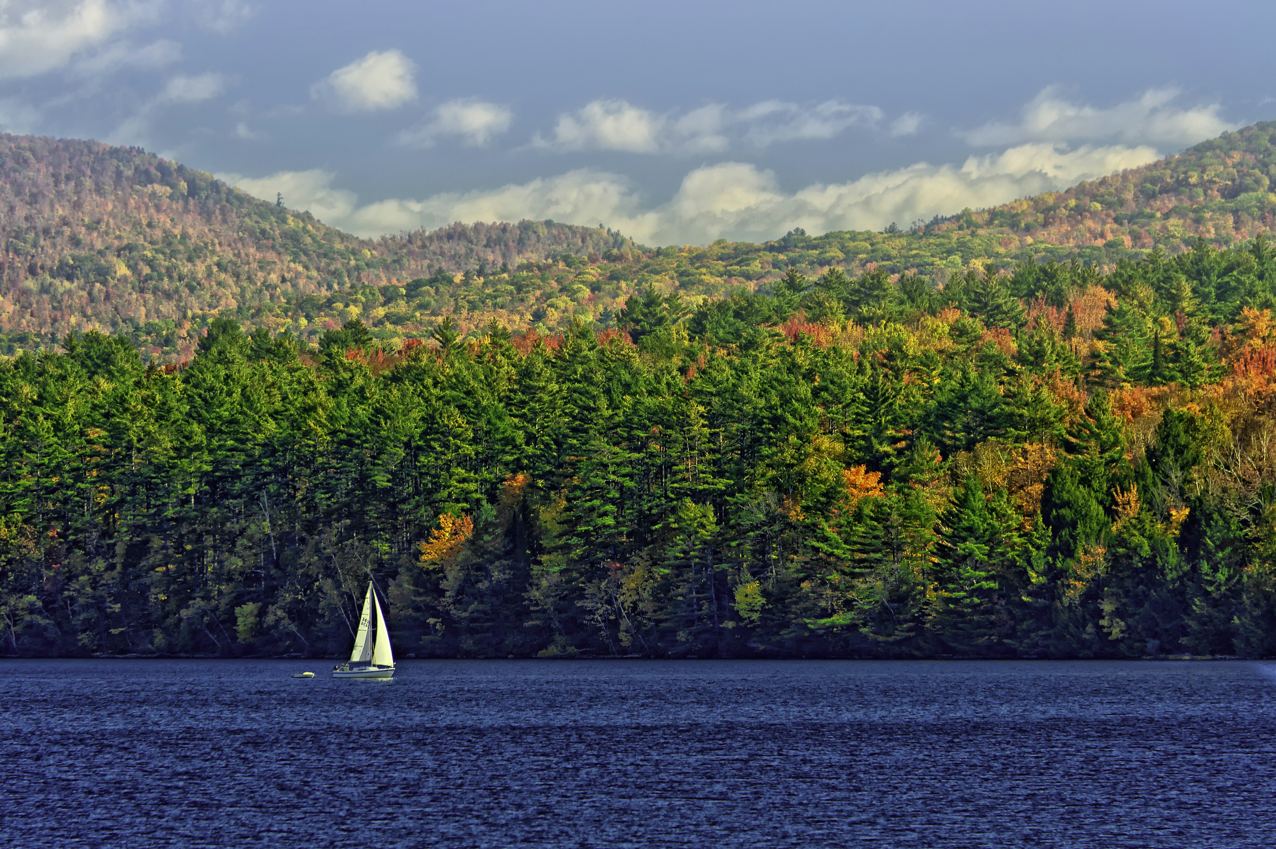 A sailboat cruises on a pristine lake as the trees on the hill behind just start to change colors; New England fall foliage road trip