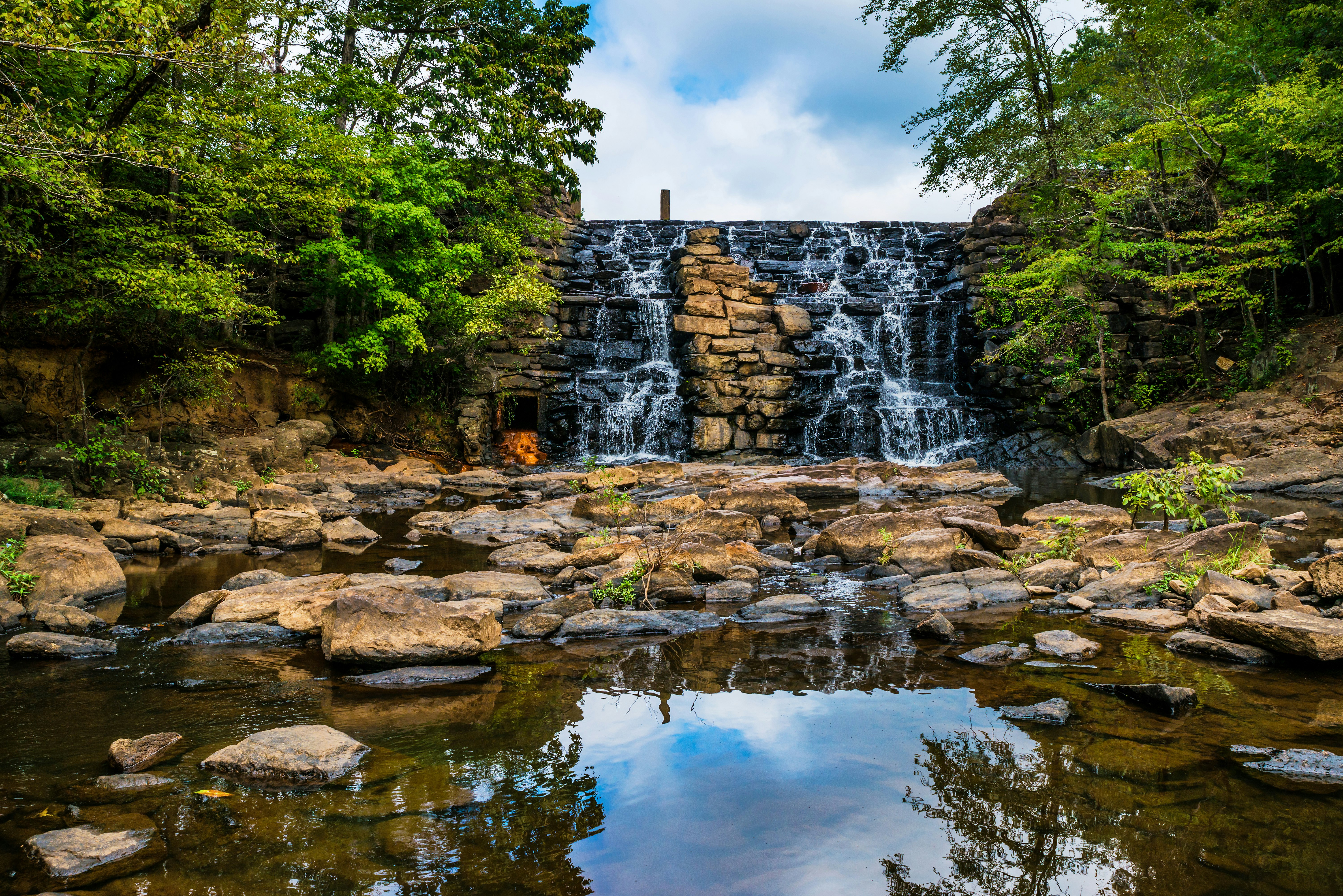 A small waterfall rushes over a stacked rocks and pools into a small pond in a wooded area; Tuscaloosa vs Auburn 