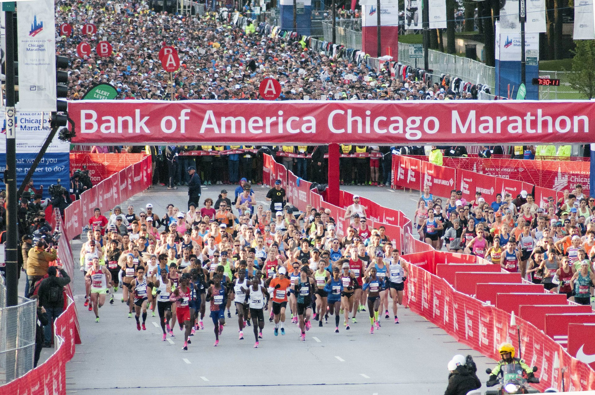 Thousands of runners begin the Chicago Marathon. There is a large red and white 'Bank of America Chicago Marathon' banner overhead.