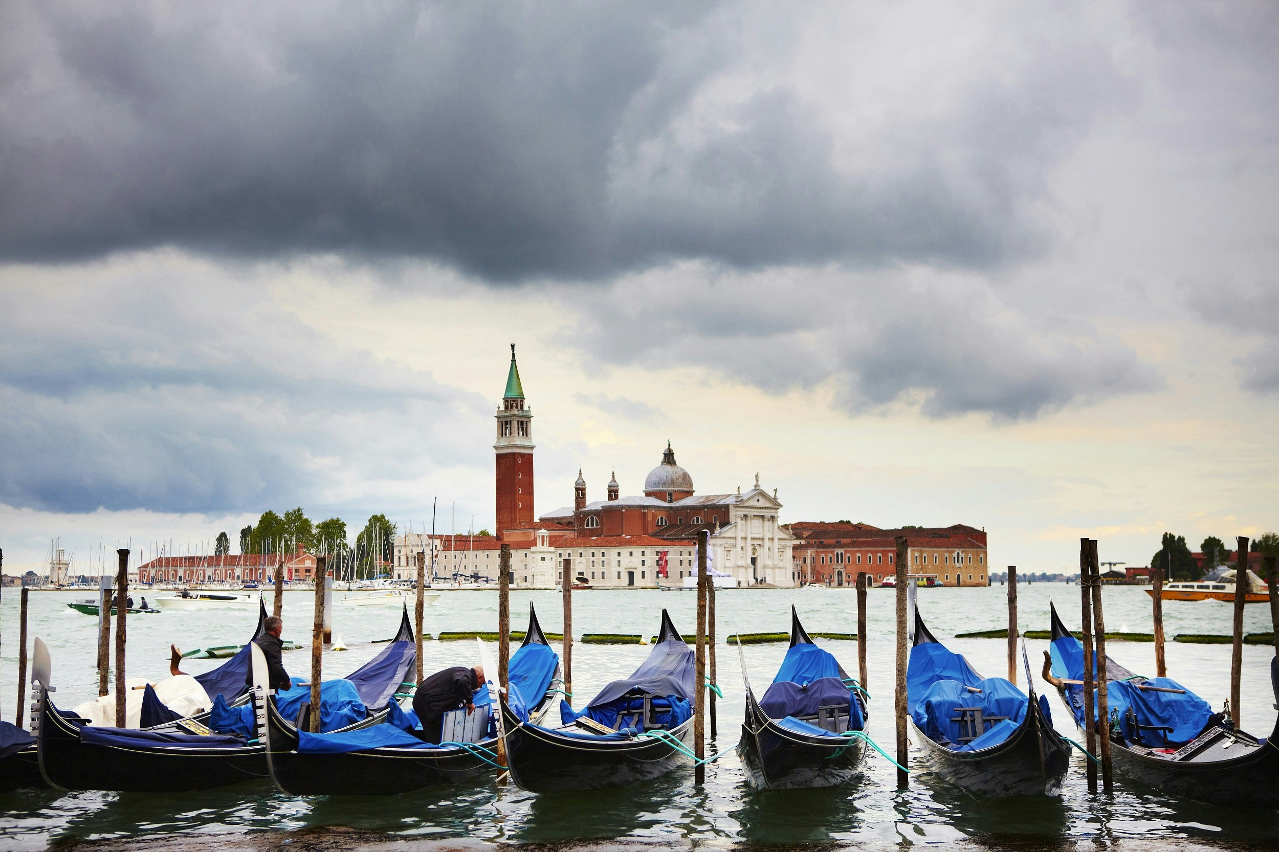 Seven gondolas with blue covers are tied to posts in the water. Across the basin is a red-brick church with a tall tower.