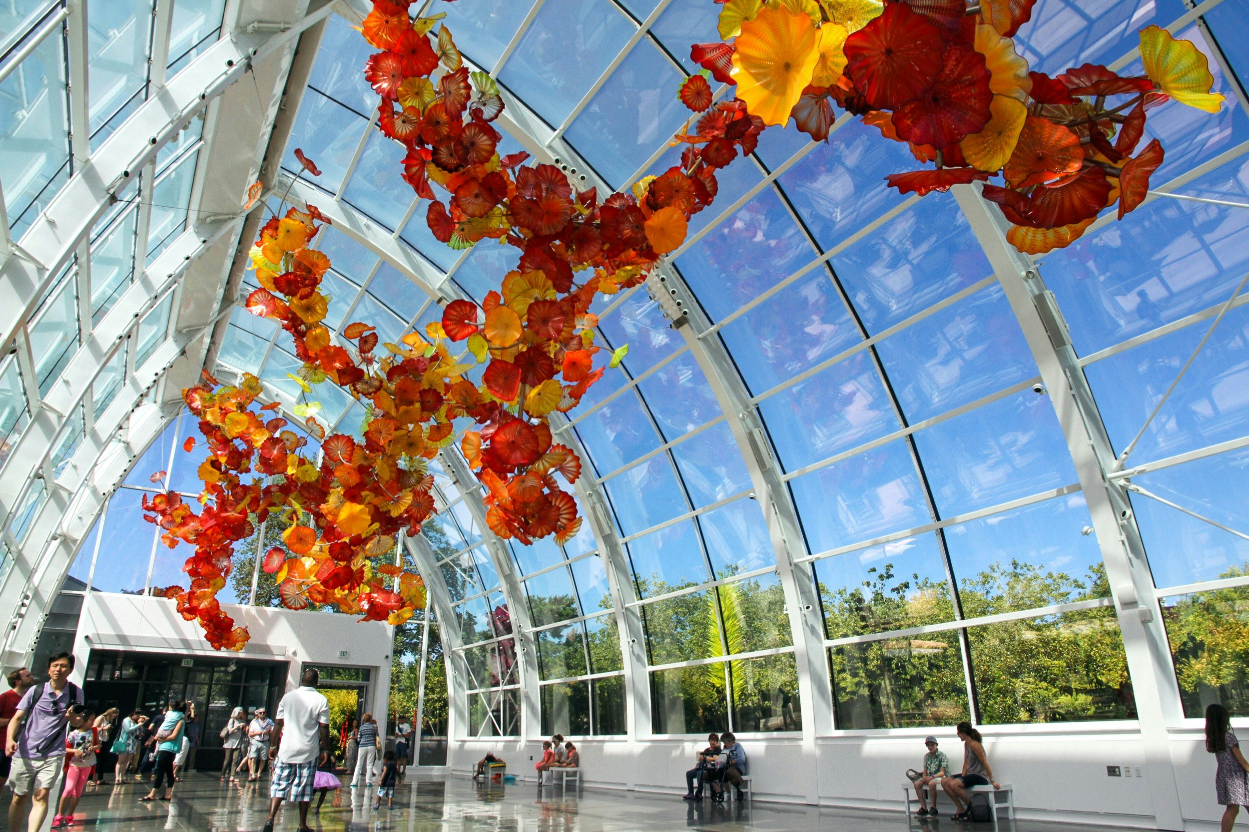 Red and orange flowers are suspended from the ceiling of a clear glass atrium as people walk below on a sunny day; Where to see Chihuly in America